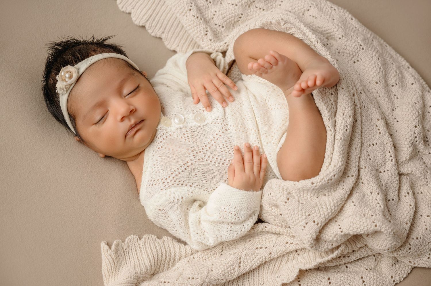 Sleeping baby wearing white knit clothing on neutral bedding.