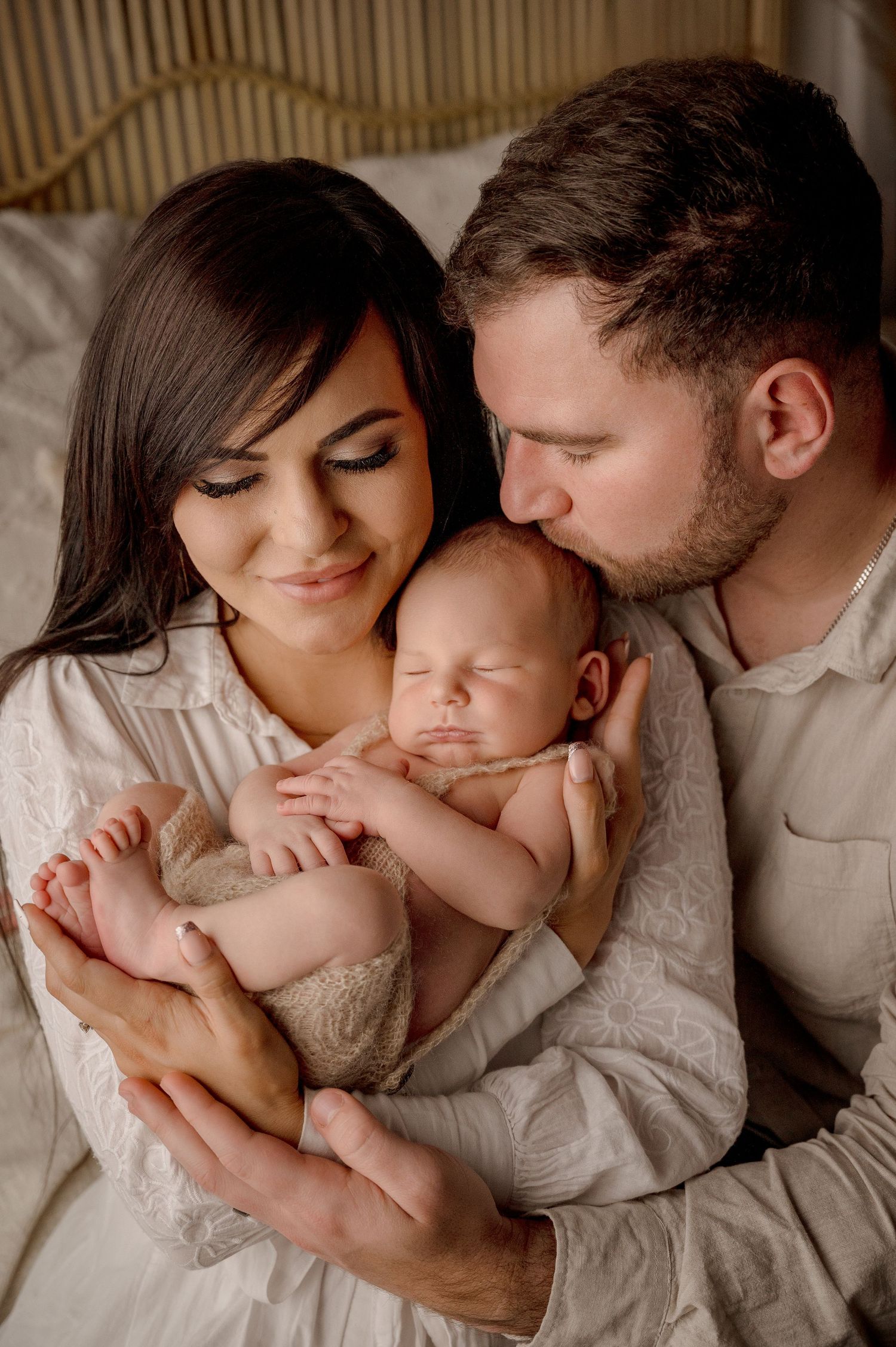 Parents lovingly embrace their newborn baby while laying together on soft white bedding in an intimate family moment.