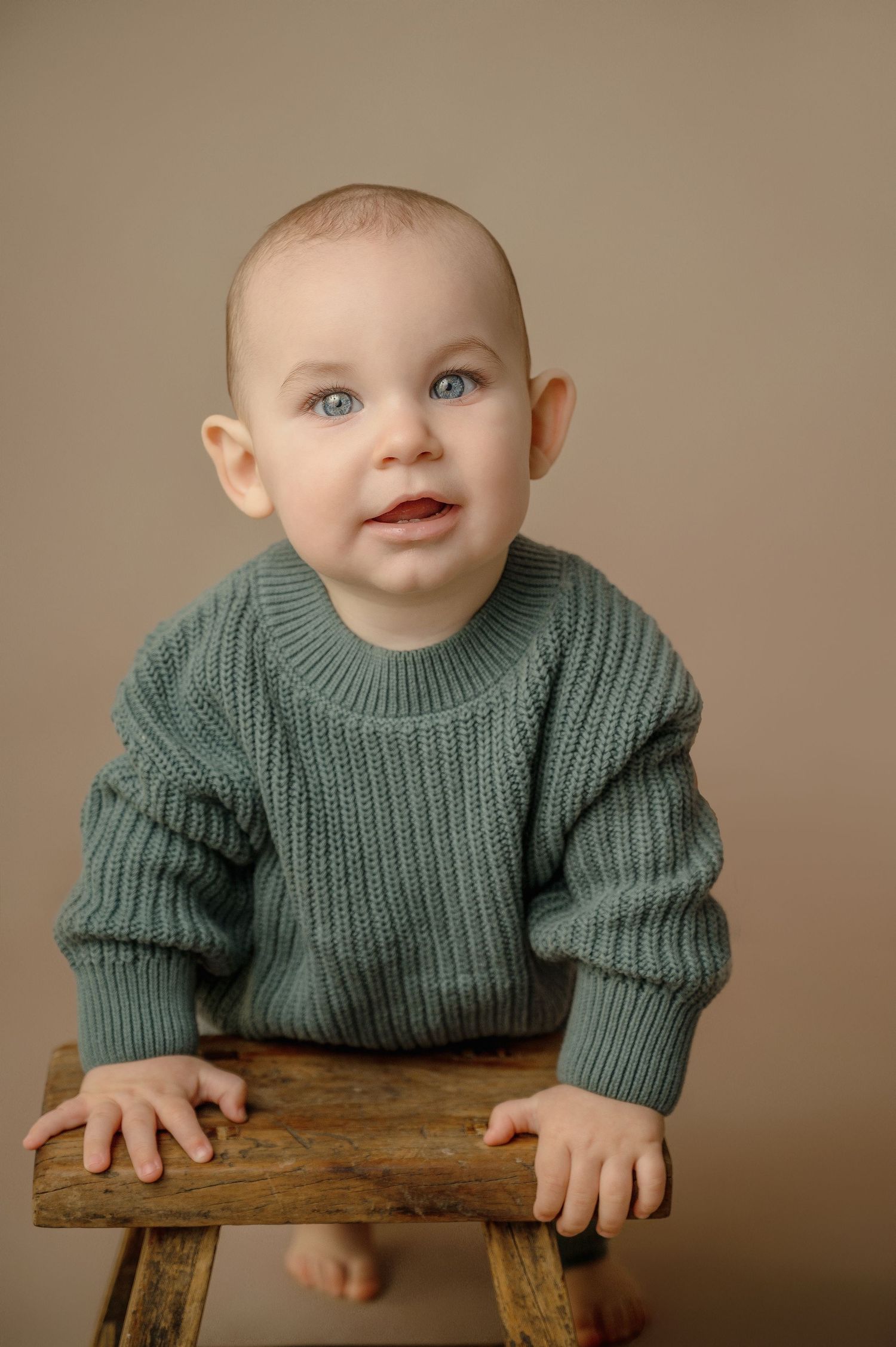 Baby in green knit sweater sits on wooden stool against neutral background.