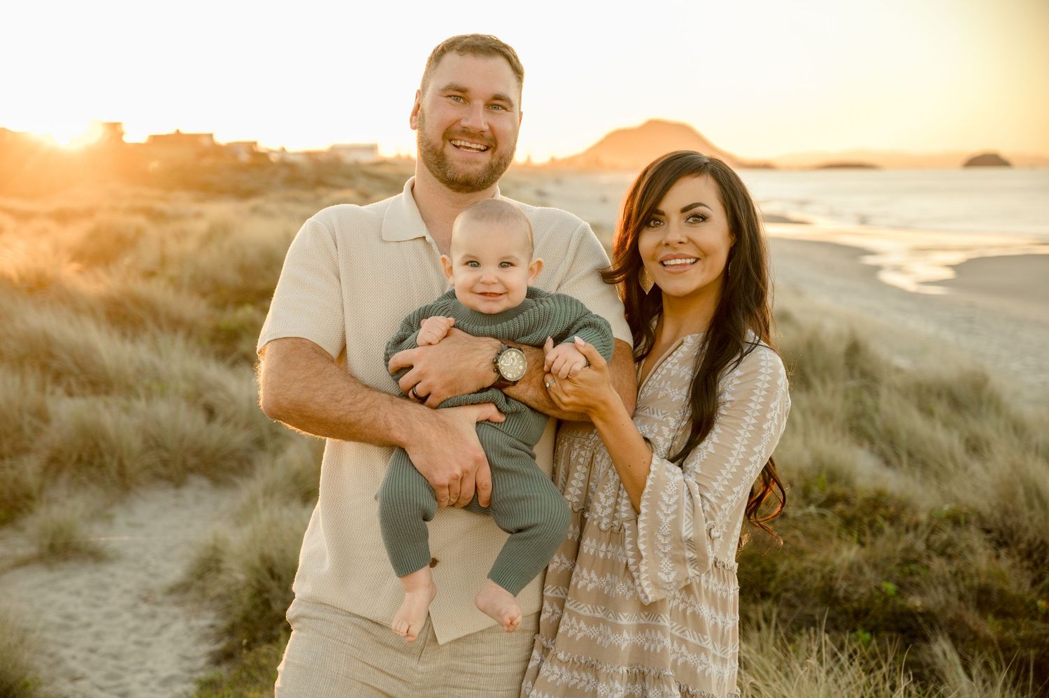Family poses together at sunset on sandy beach dunes with ocean in background.