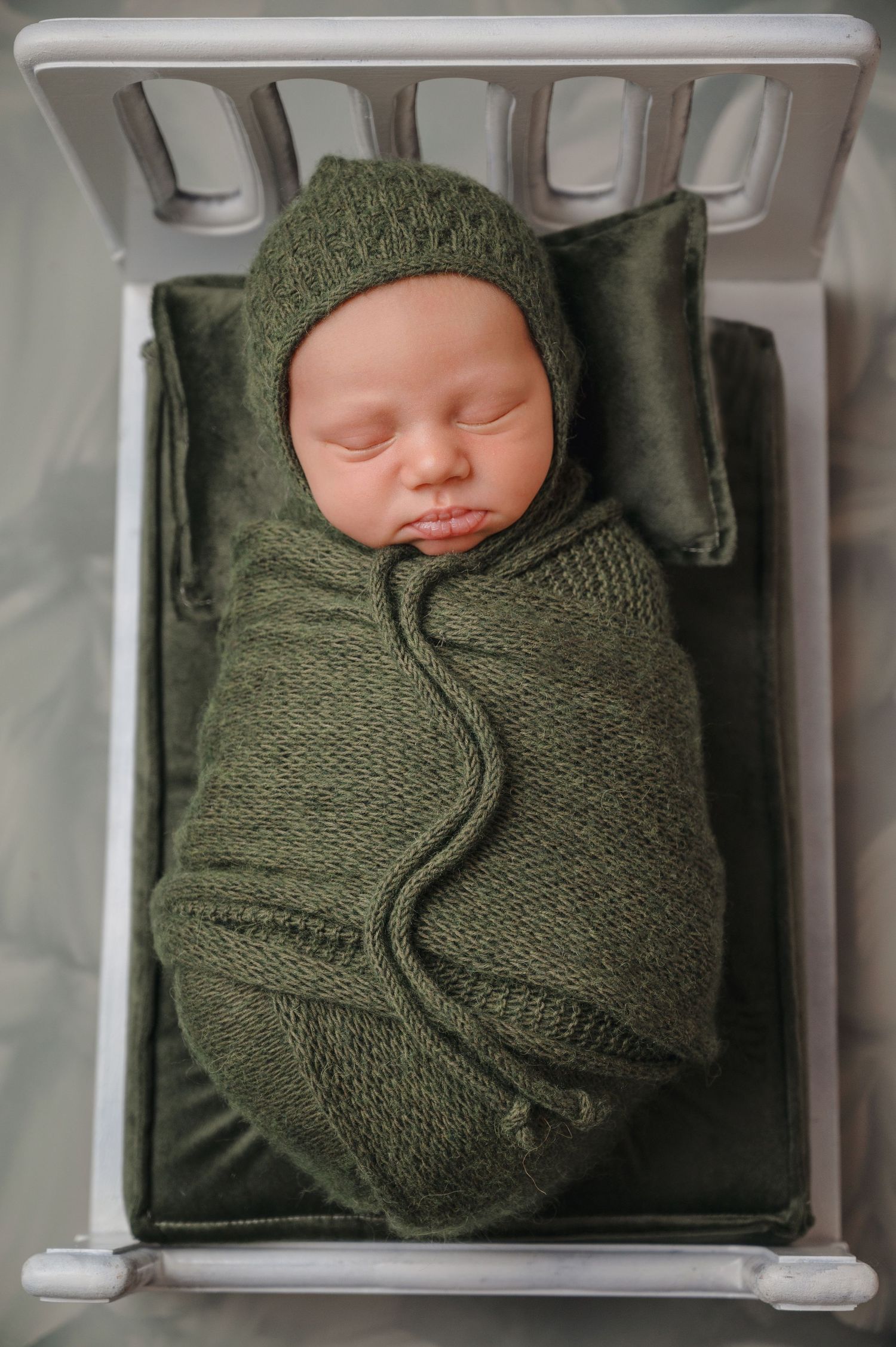 A sleeping newborn wrapped in olive green knitted swaddle and matching hat lies on a cushioned surface.