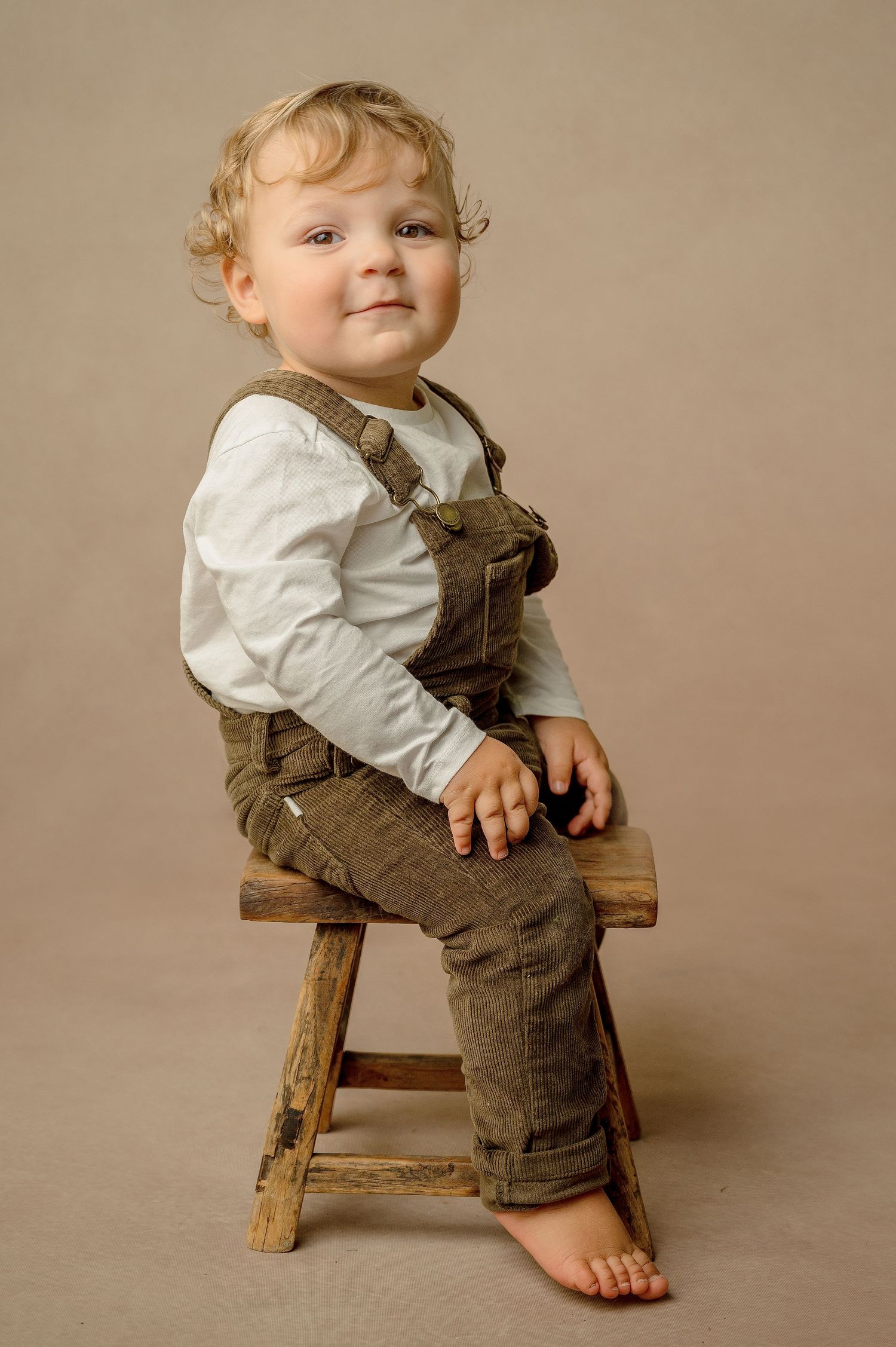 A toddler wearing brown overalls and a white shirt sits on a wooden stool against a beige background.
