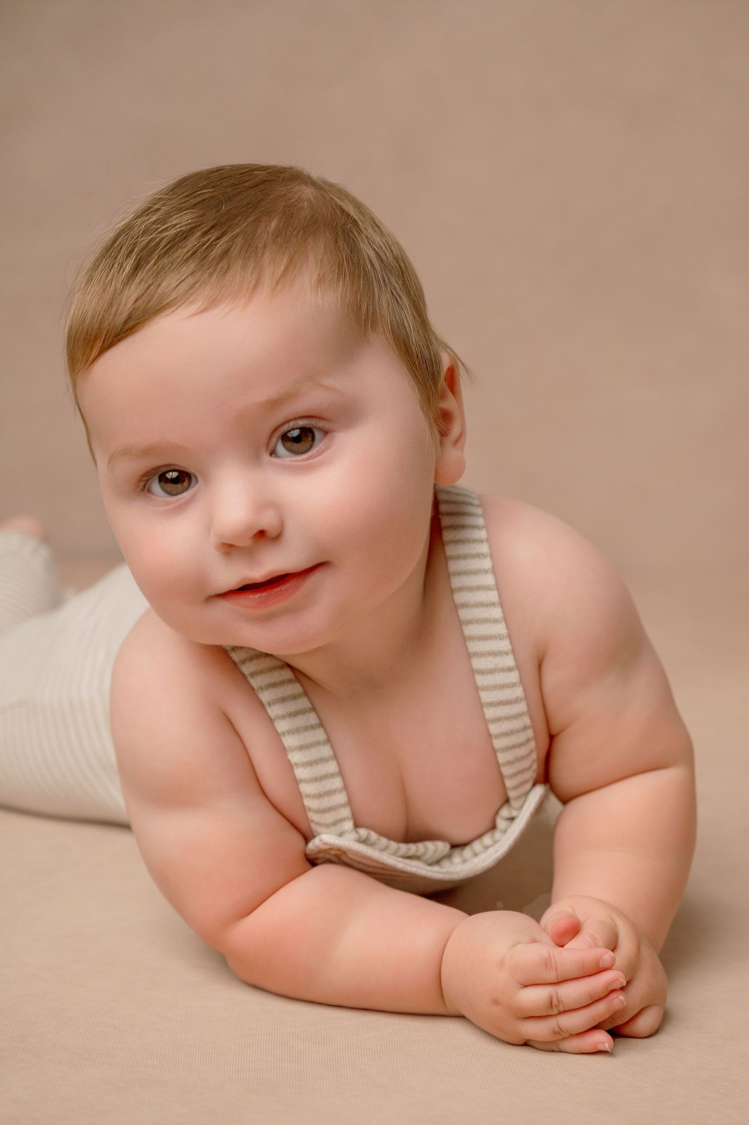A baby in striped overalls lies on their stomach on a beige backdrop during a studio photoshoot.
