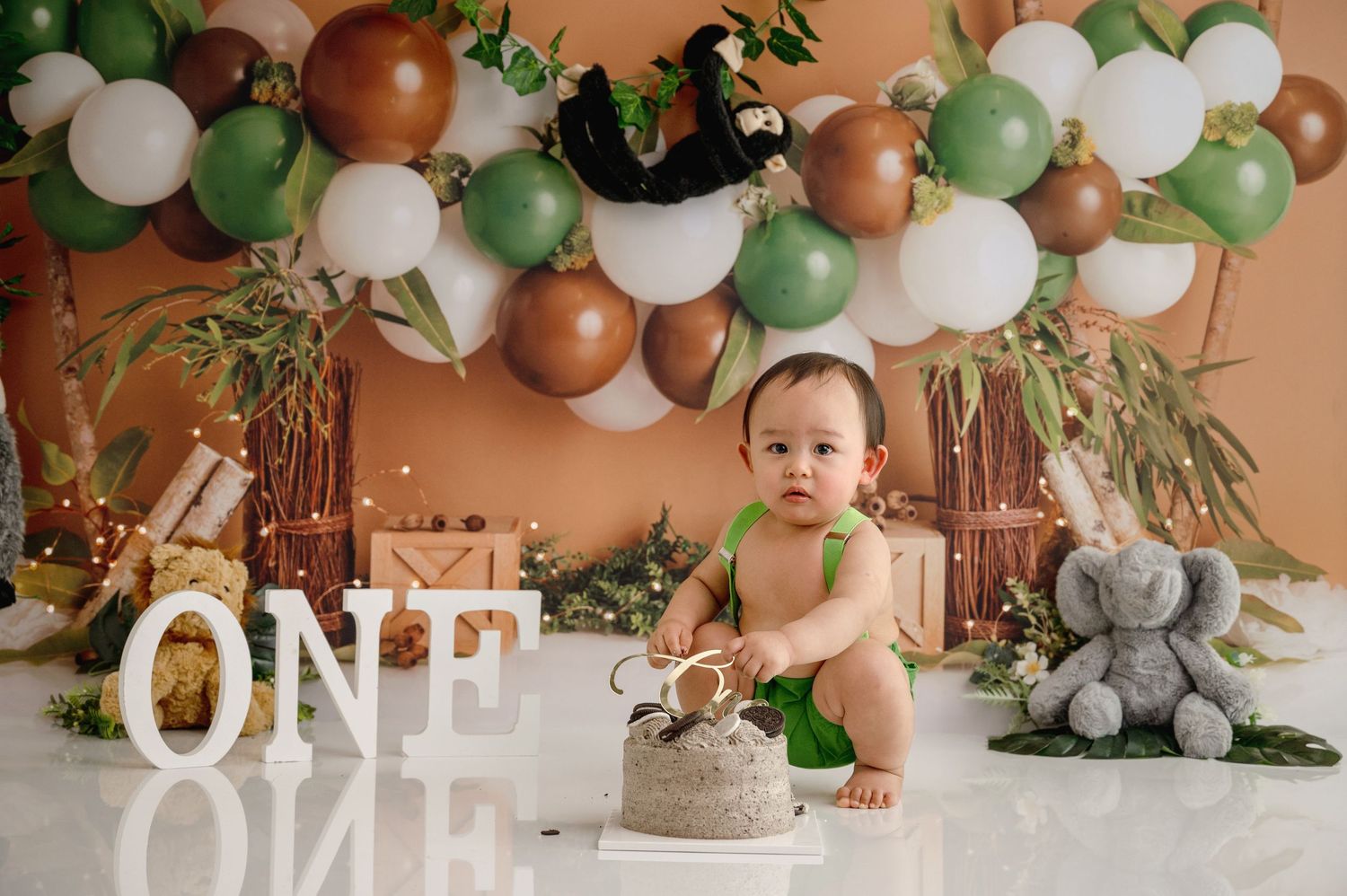 A first birthday setup with balloon arch, cake, and decorative ONE letters against coral background.