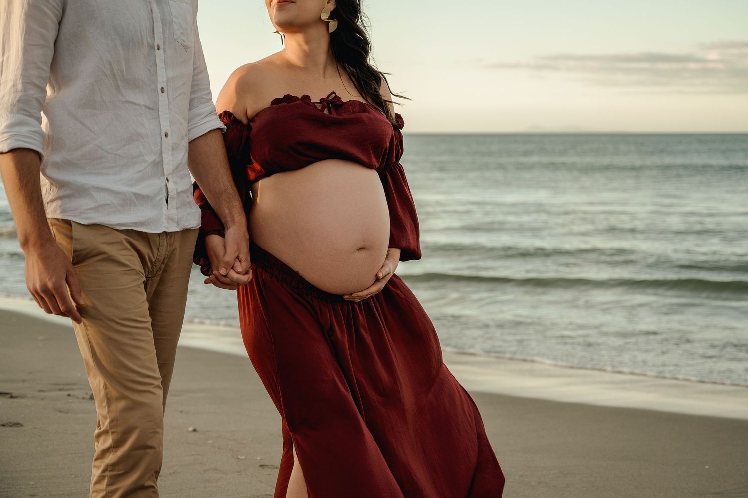 A romantic photoshoot at sunset on a beach with a couple in burgundy dress and khaki pants walking in the sand.