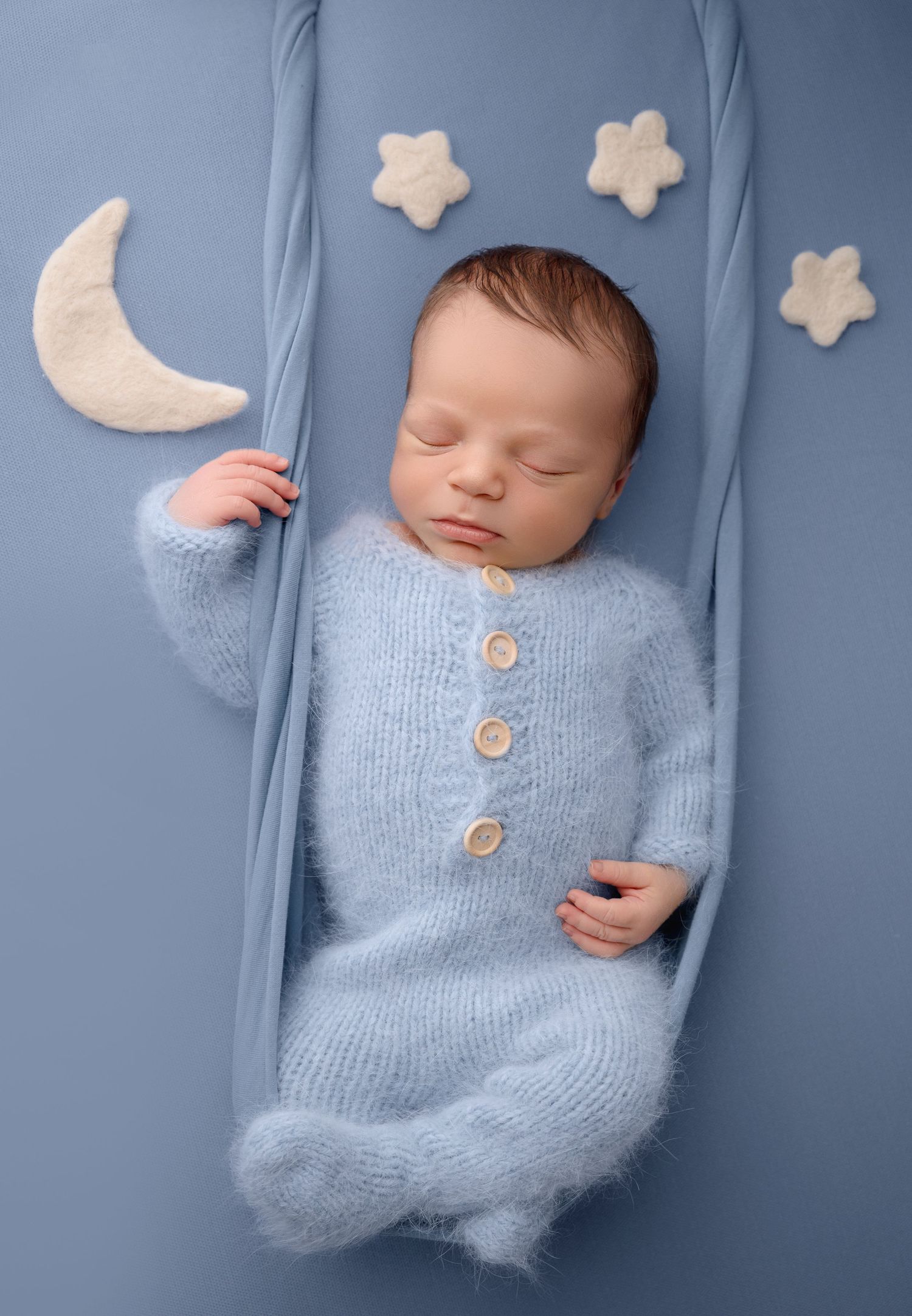 A sleeping baby in a light blue knit romper lies on a blue backdrop with decorative stars and moons.