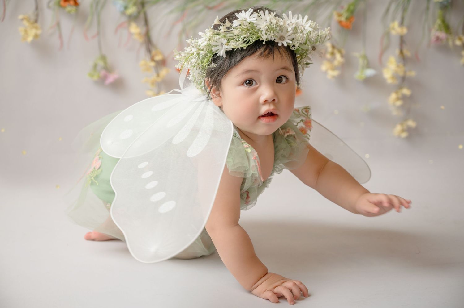 A baby in a white dress and floral crown crawls on a white floor with hanging flowers in the background.