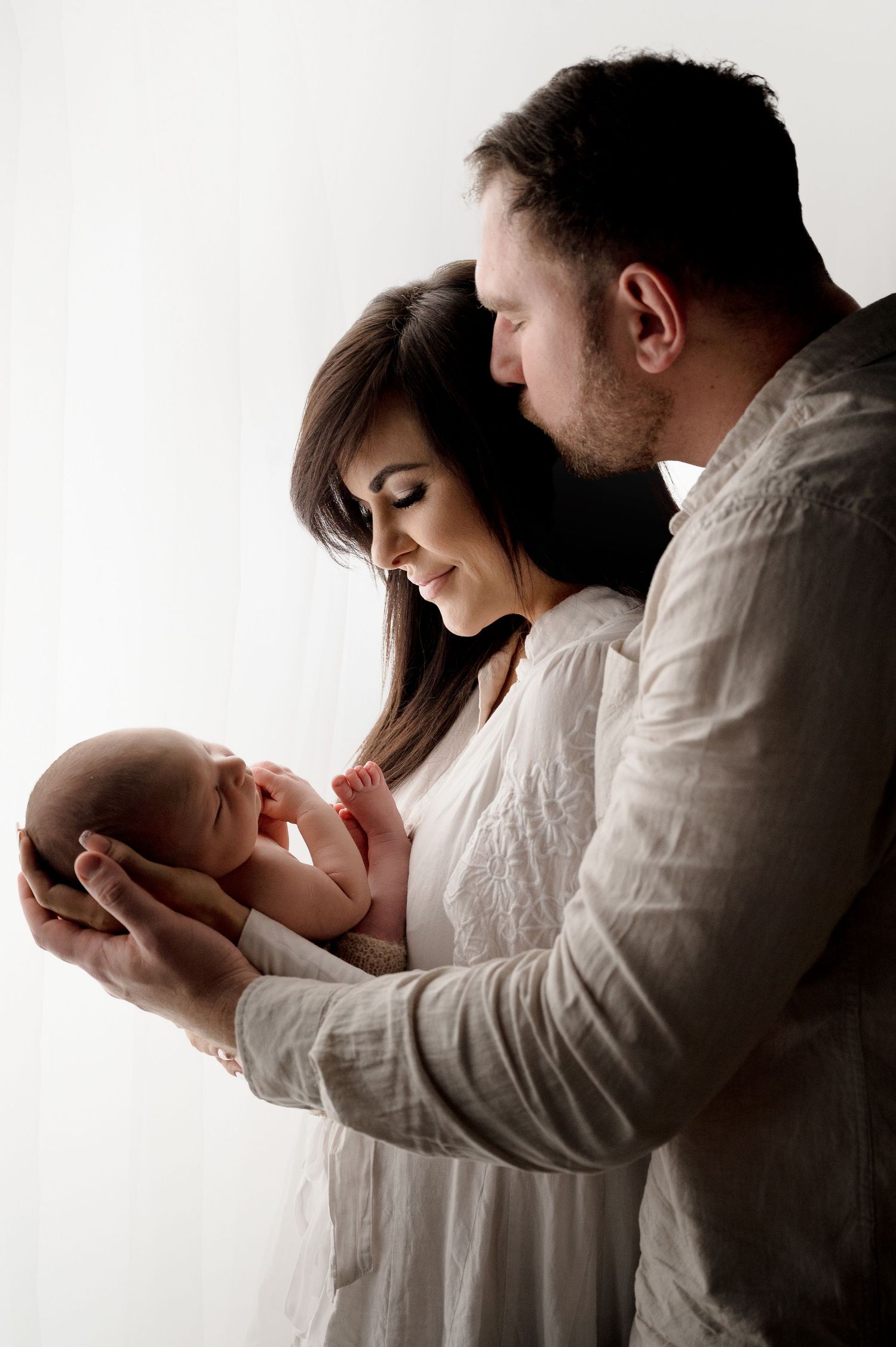 A couple in white clothing gently hold and look lovingly at their newborn baby against a bright background.
