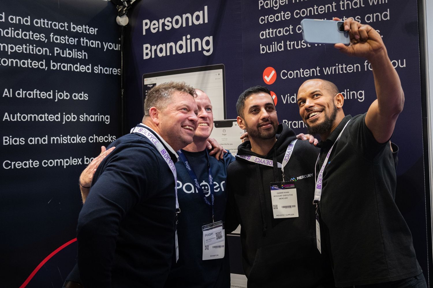 Group of conference attendees taking selfie at exhibition booth with branding backdrop.