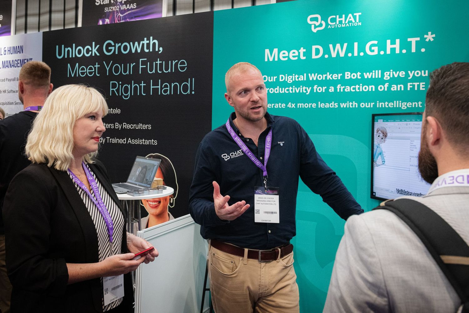 Trade show exhibitors demonstrating products at conference booth with digital marketing displays.