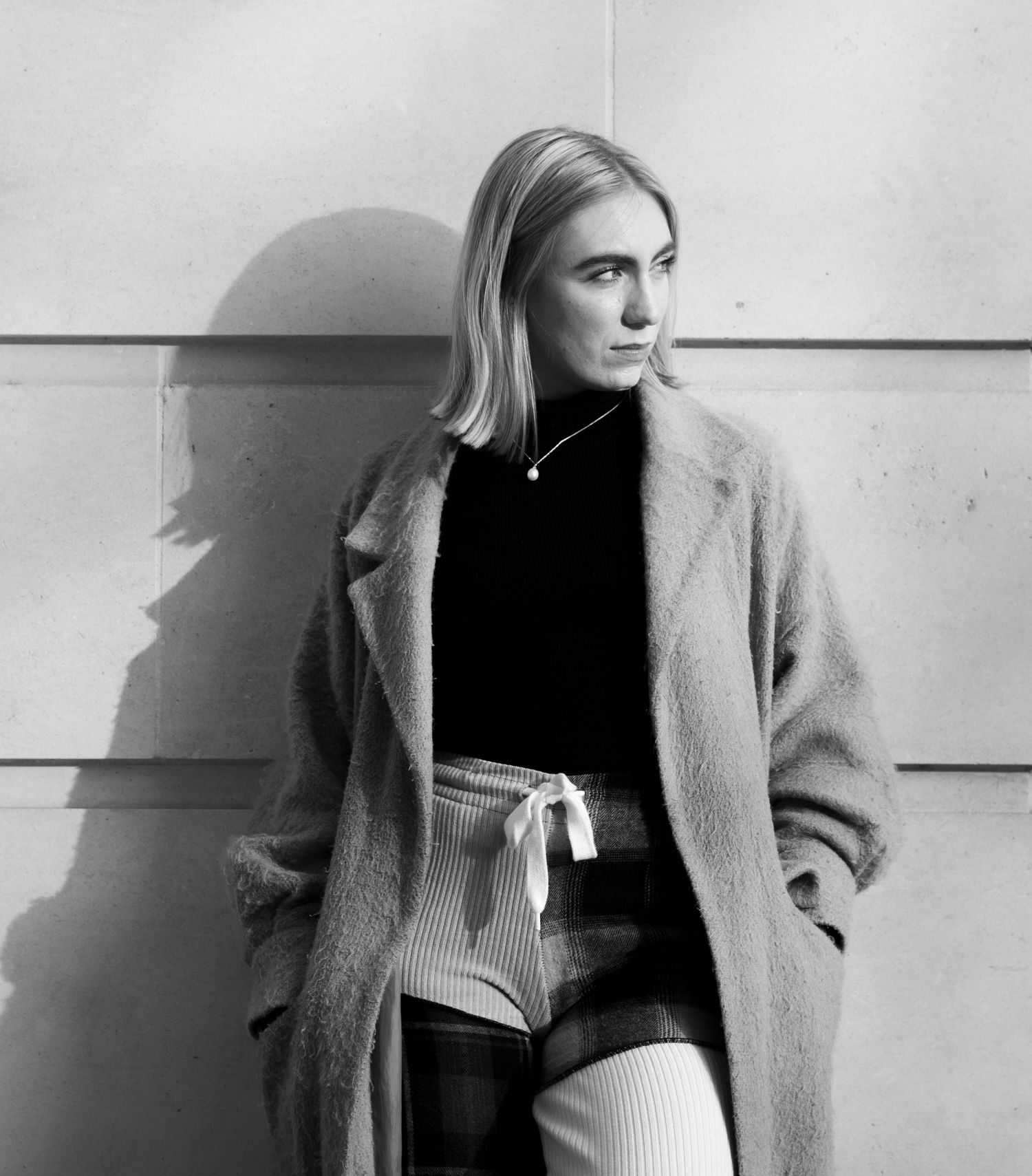 Black and white portrait of someone in winter coat leaning against wall with dramatic lighting.