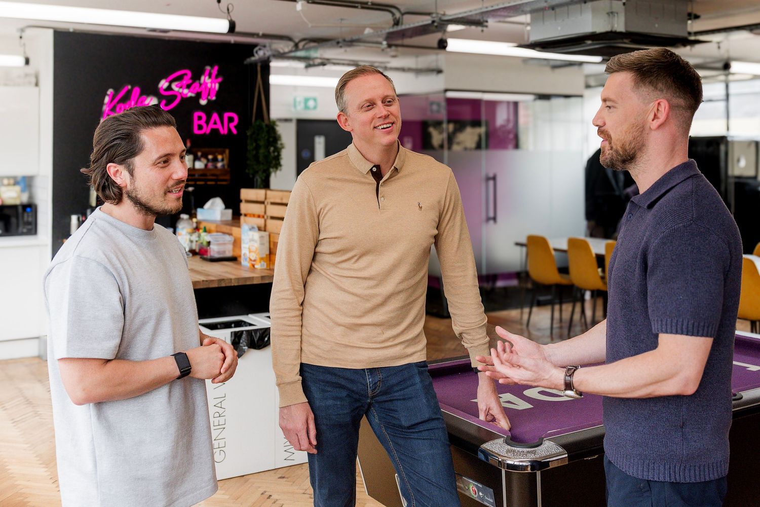 Three colleagues engaged in discussion at a modern office space with pink neon signage.