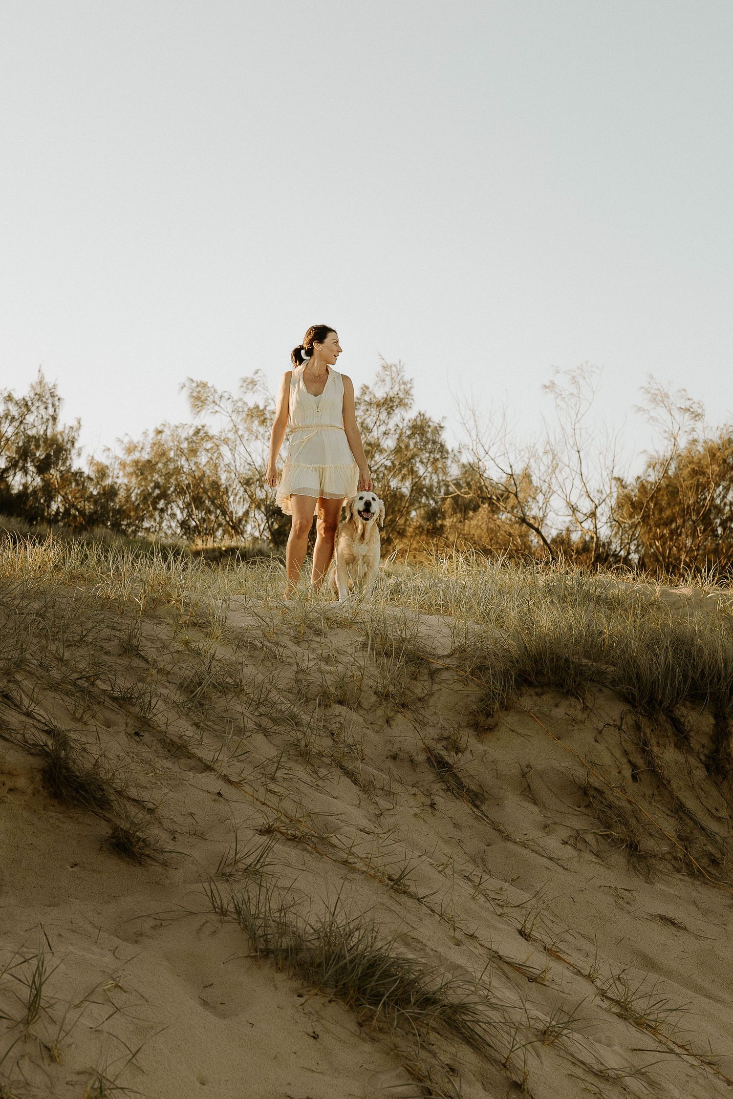 A figure in a white dress stands on a sandy dune at golden hour as sunlight casts long shadows across the landscape.