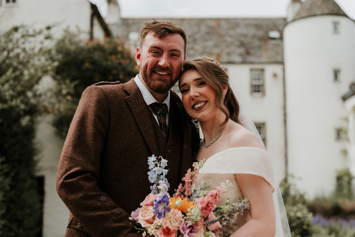 A couple shares a joyful moment at their wedding with a colorful bouquet in front of a historic stone castle.