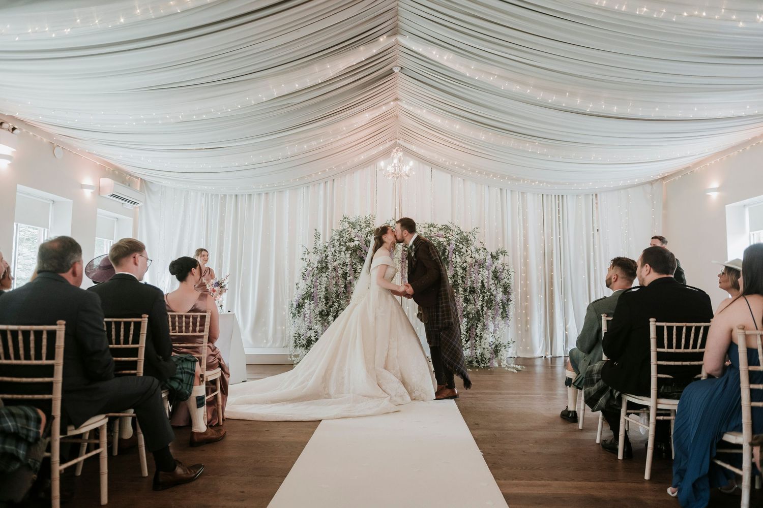 An elegant indoor wedding ceremony in a white draped tent with guests seated on chiavari chairs and a floral arch backdrop.