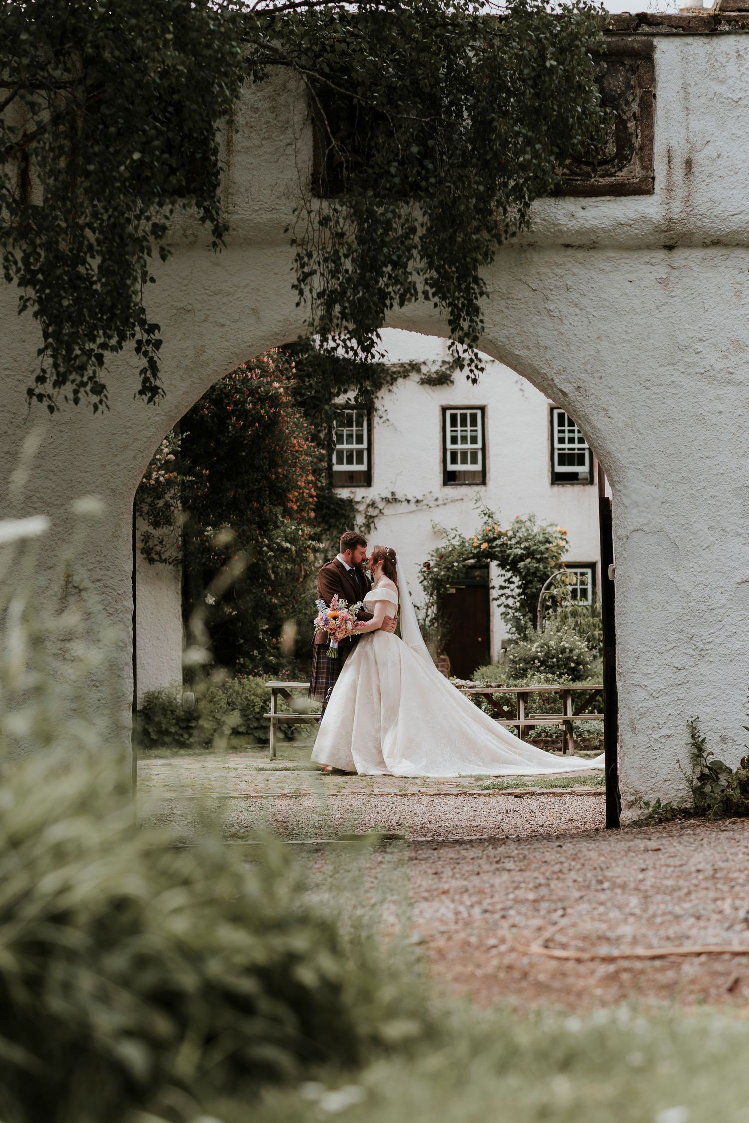 Romantic wedding couple embraces under stone archway framed by greenery with white colonial style building in background.