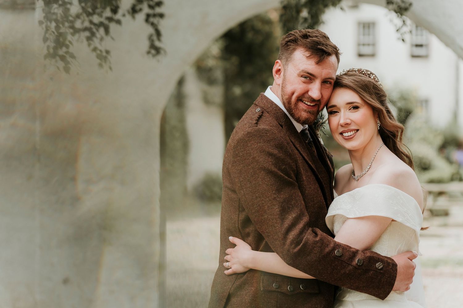 A couple embraces and smiles outdoors under a stone archway with soft romantic lighting.