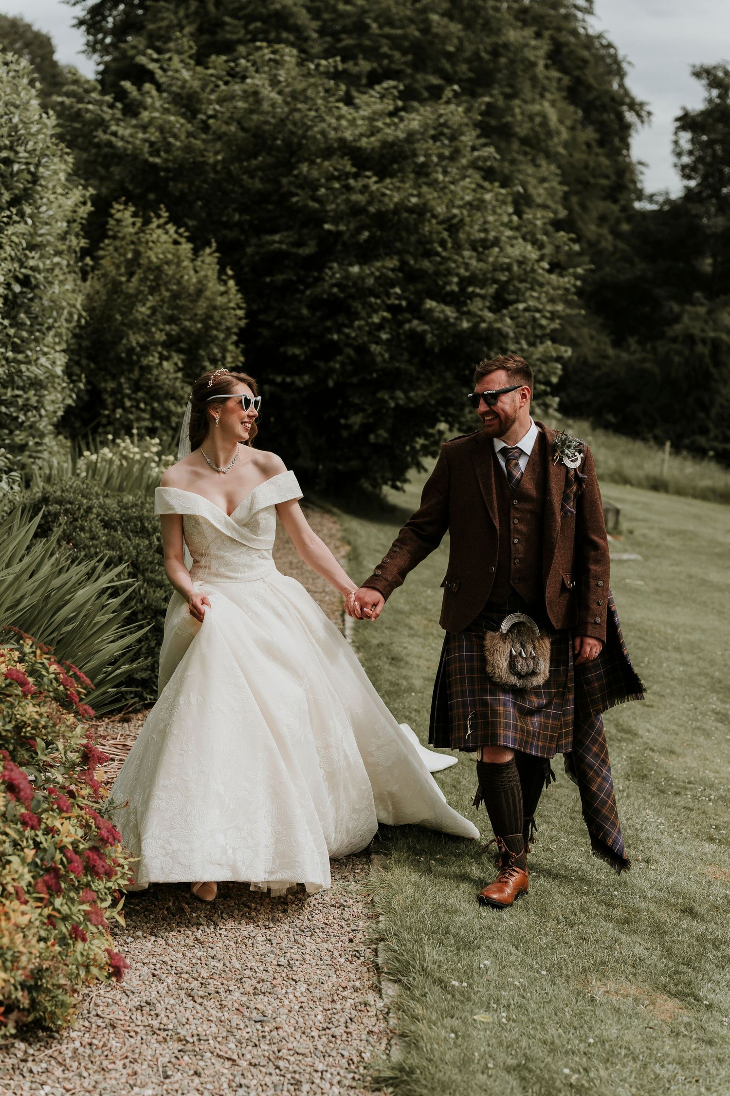 A bride in a flowing white ballgown and groom in traditional Scottish attire walk together on a grassy path.