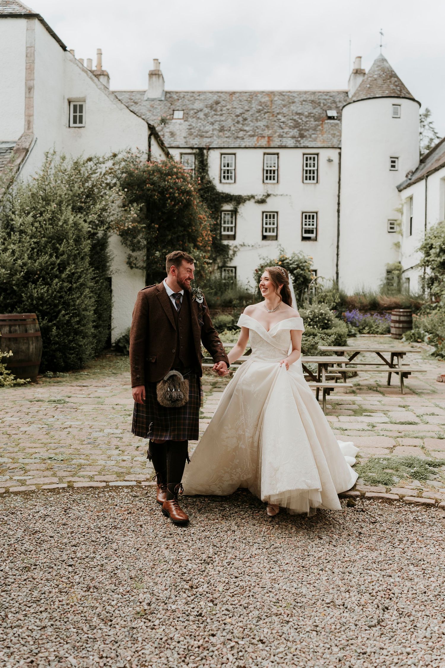 A wedding photoshoot on a cobblestone path outside a traditional Scottish stone manor with cream-colored walls.