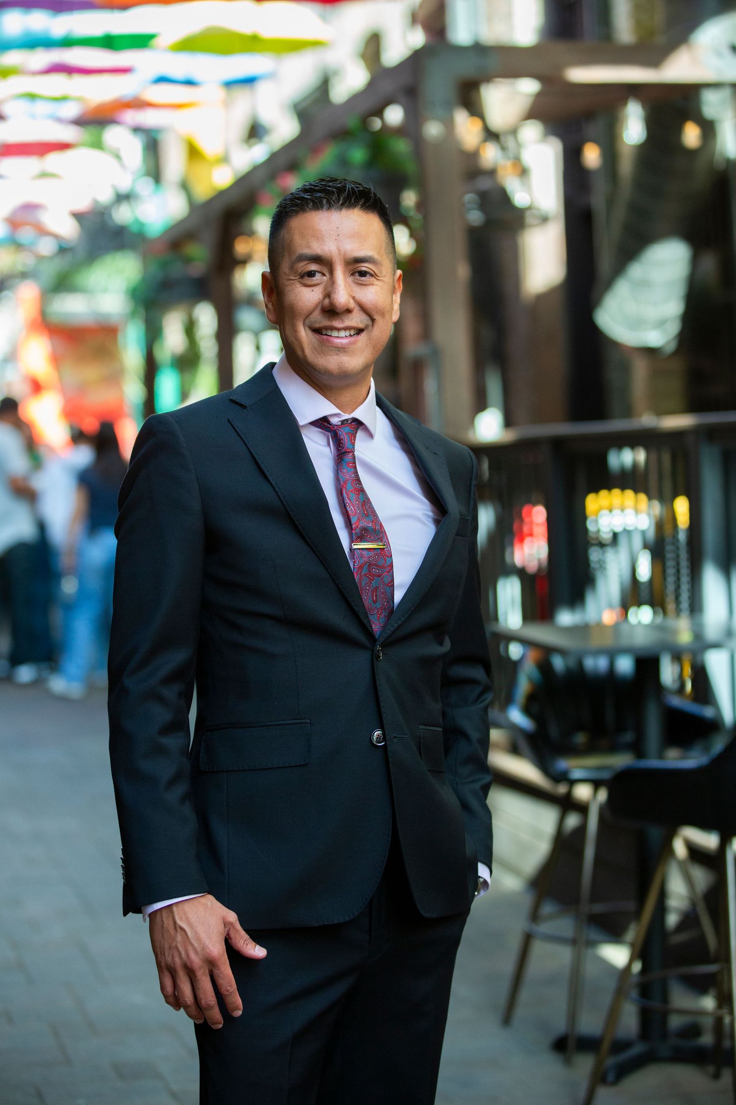 Professional in dark suit and burgundy tie standing on vibrant urban street with colorful decorations overhead.