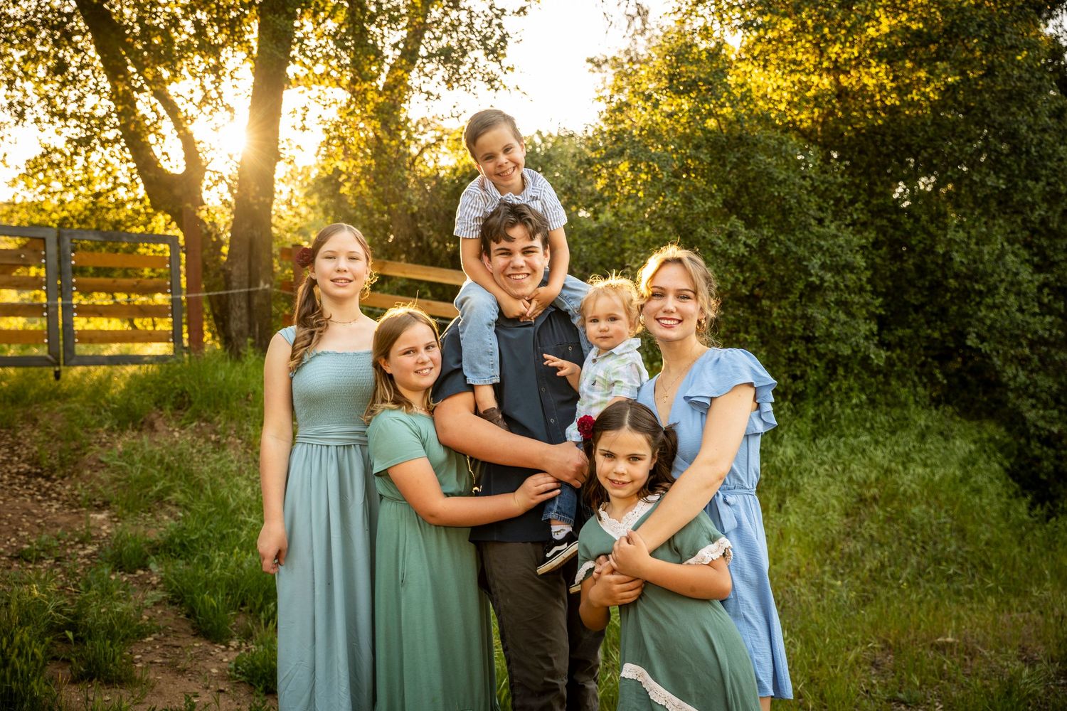 A group in sage green dresses and casual attire poses together at sunset in a wooded outdoor setting.