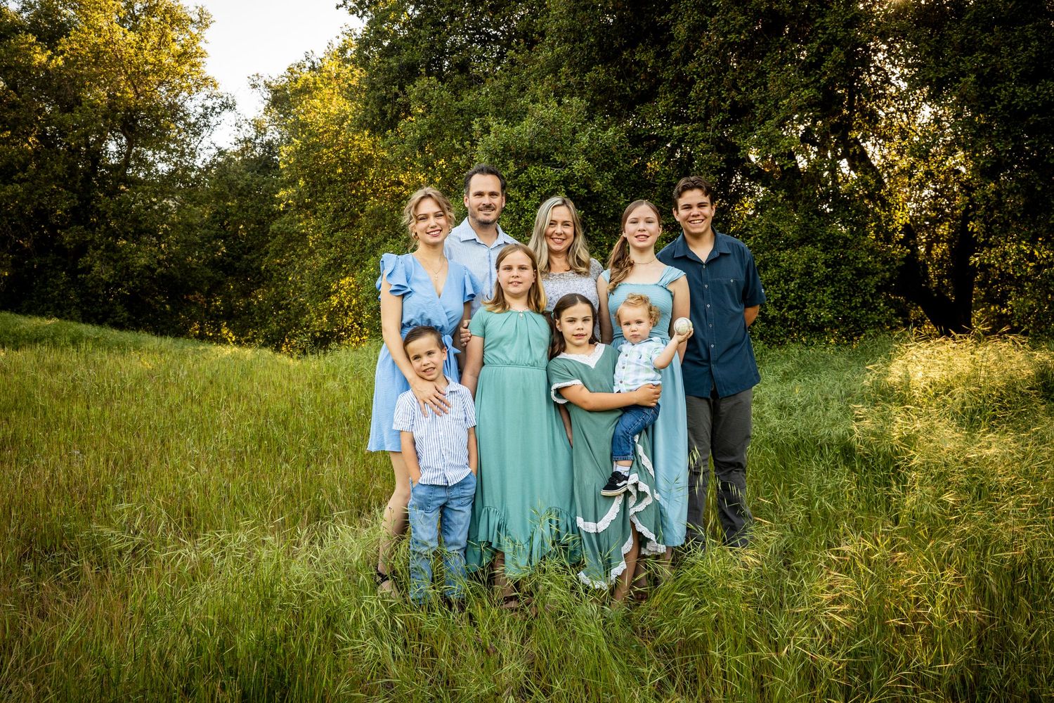 Group wearing coordinated blue and turquoise outfits pose together on a grassy hillside during golden hour.