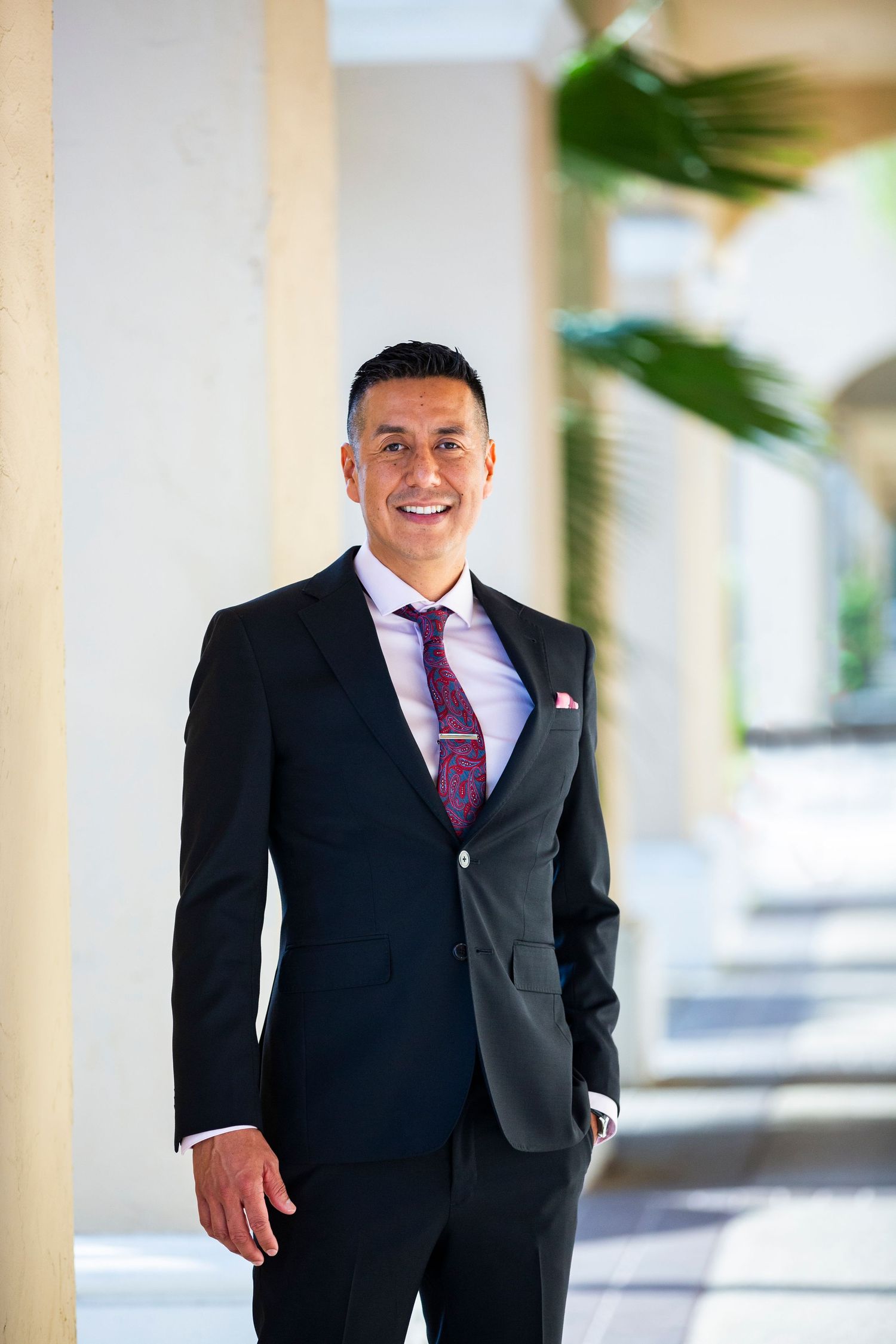 Professional in a black suit and purple tie walks confidently through a bright corridor with pillars.