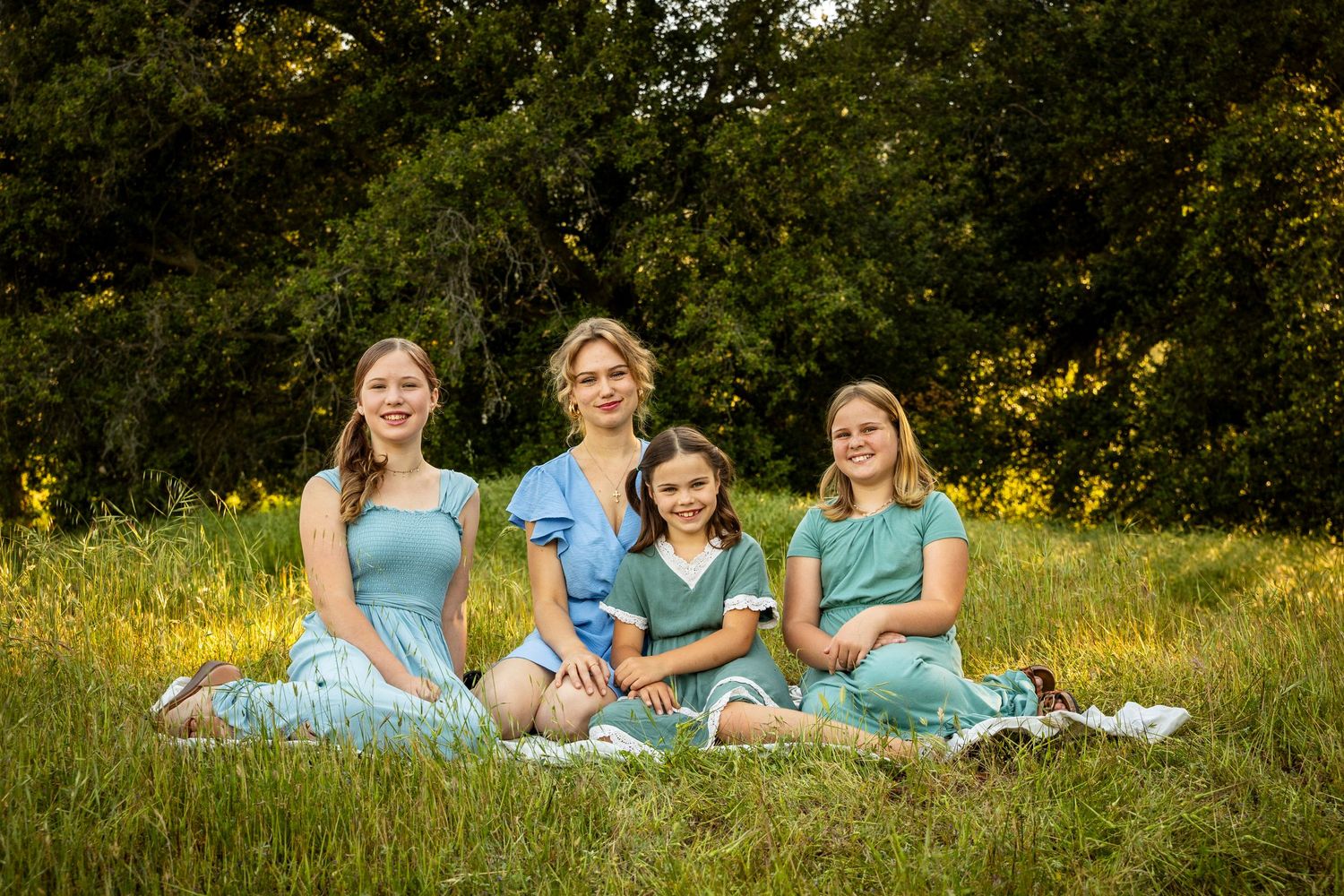 Four people in matching mint blue dresses sit together on grass in a sunny outdoor setting.