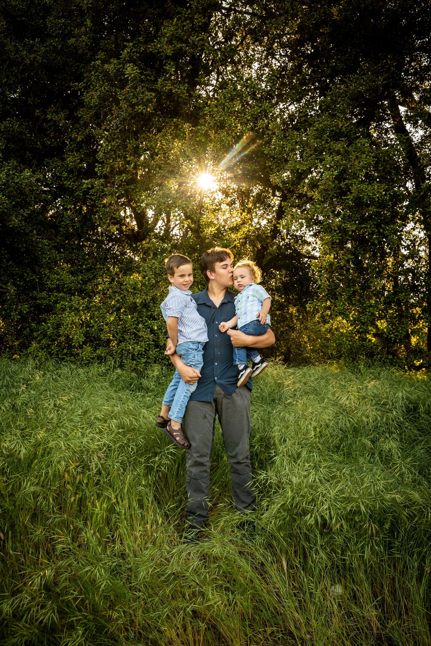 A parent and children pose in a grassy field at sunset with sunbeams bursting through tree leaves in the background.