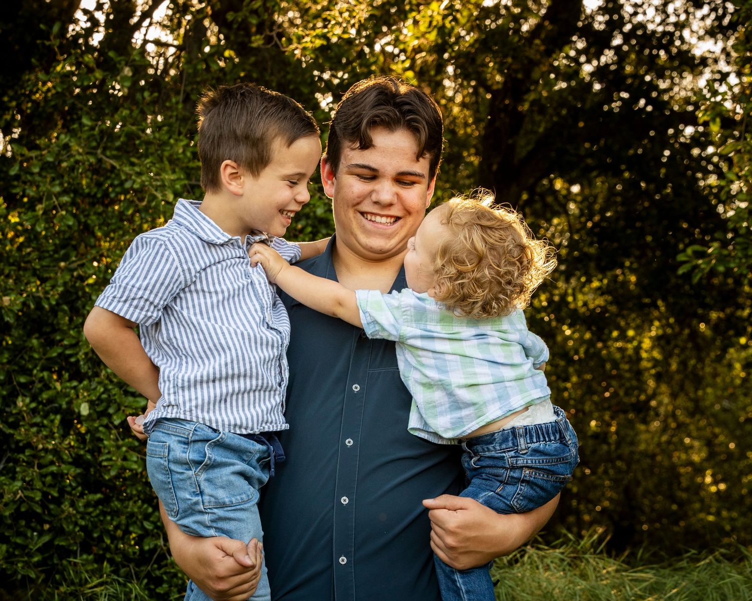 Three people hugging and smiling together outdoors during golden hour in a wooded setting.