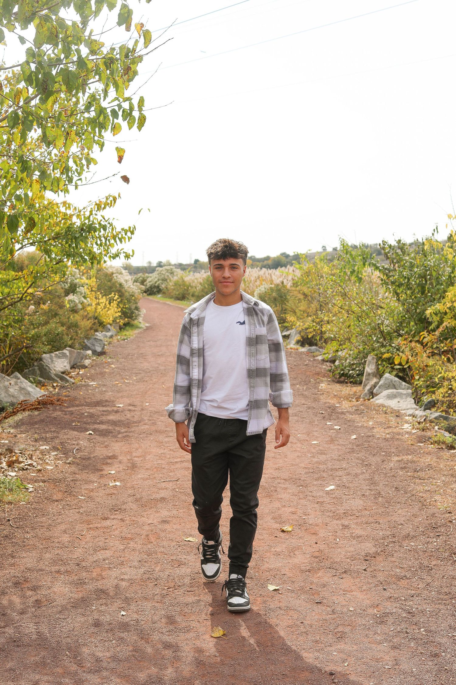 Student in plaid shirt and white tee walks along dirt path with autumn trees.
