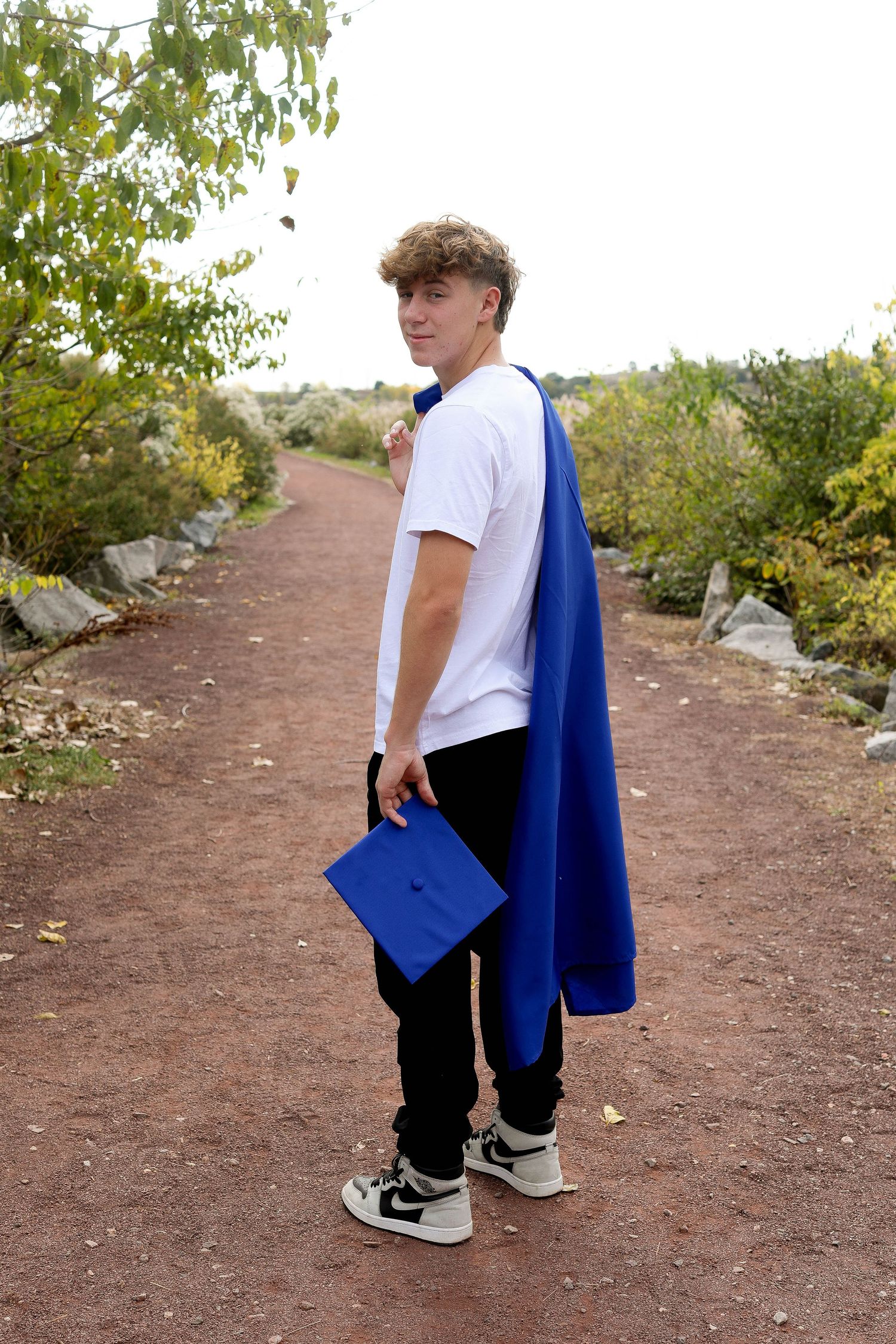 Graduate holding blue cap and gown looks over shoulder while standing on nature trail.