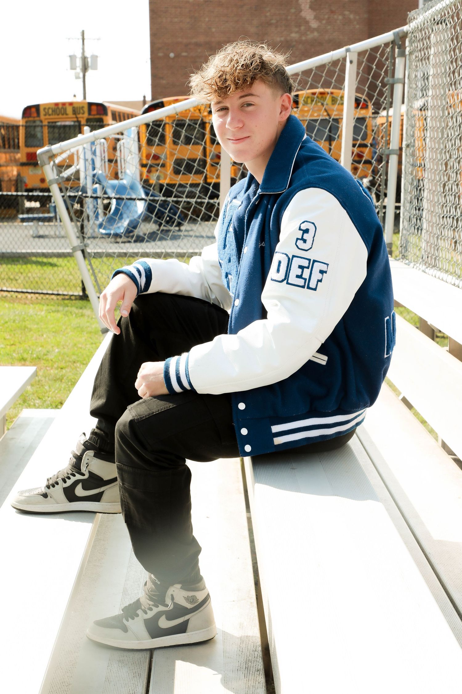 Athletic scene with someone in varsity jacket sitting on bleachers near school buses.