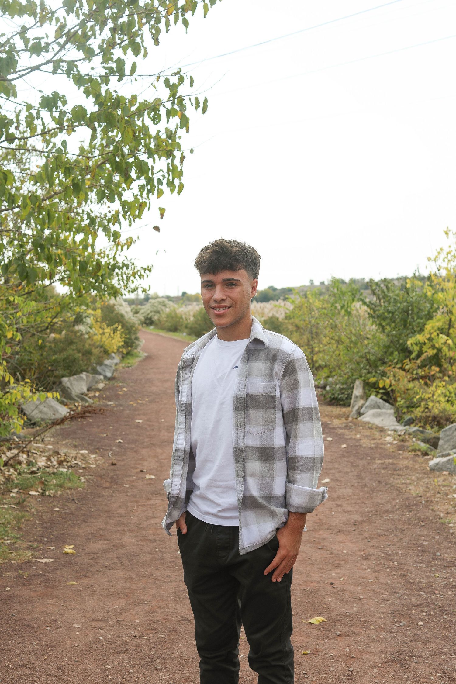 Student wearing gray plaid shirt poses on nature trail with trees in background.