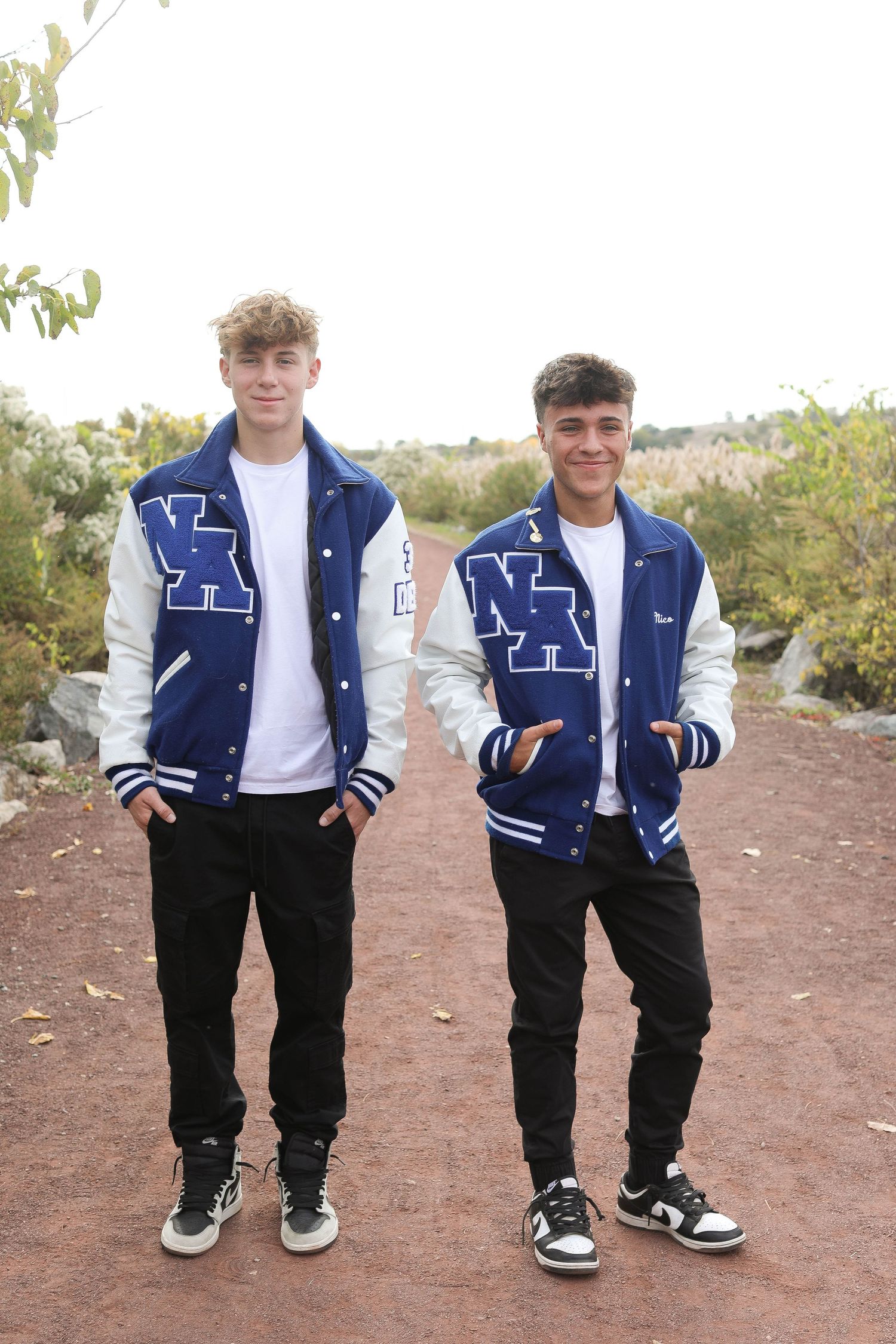 Two students pose in matching blue and white varsity letterman jackets on a dirt path outdoors.