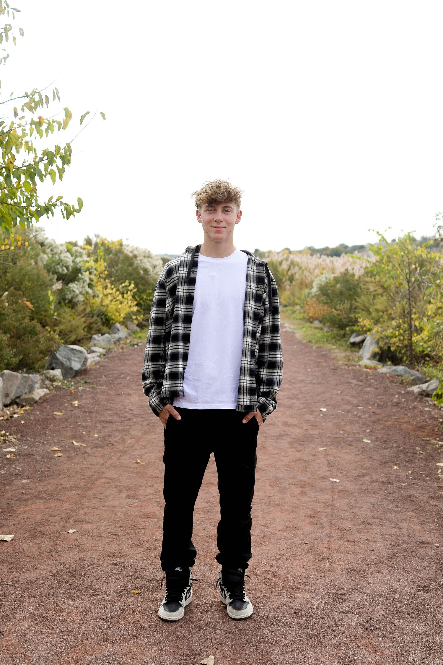 Someone poses in black and white plaid flannel and jeans on a dirt path lined with rocks and trees.