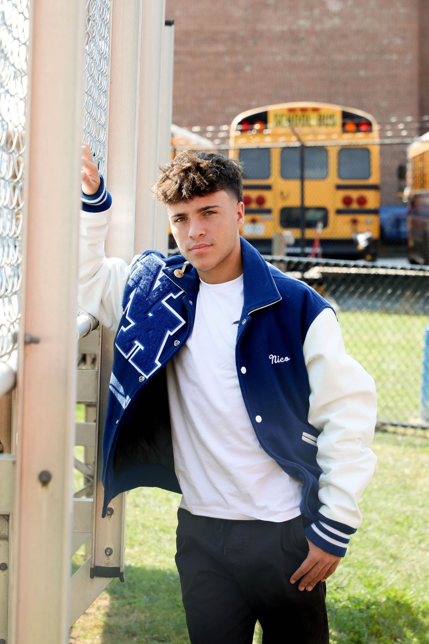 Casual pose against fence post wearing varsity jacket with school buses in background.