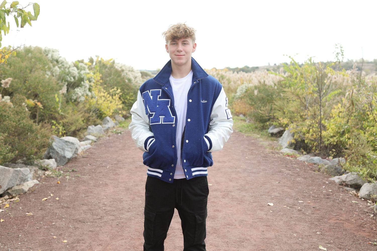 Student in blue and white letterman jacket stands on rustic path with rocks and foliage.