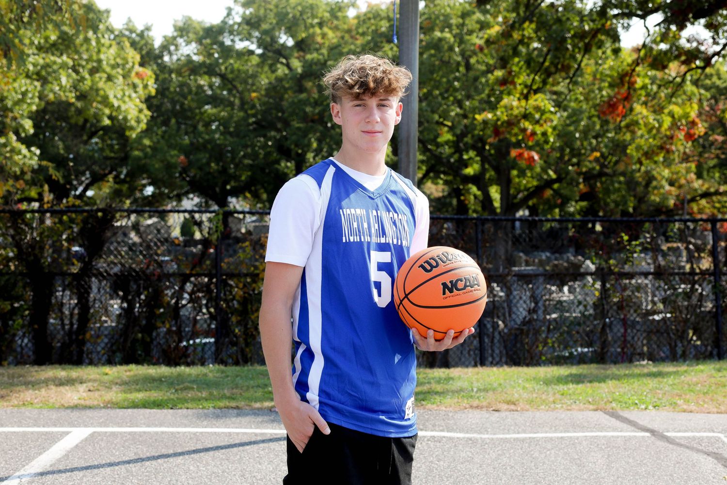 Basketball player poses with ball on outdoor court wearing blue Vikings jersey.