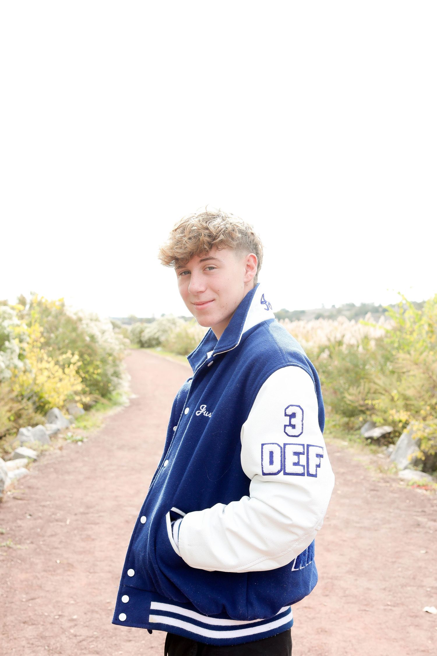 Student wearing varsity jacket with 'DEF' lettering poses on dirt trail in natural setting.