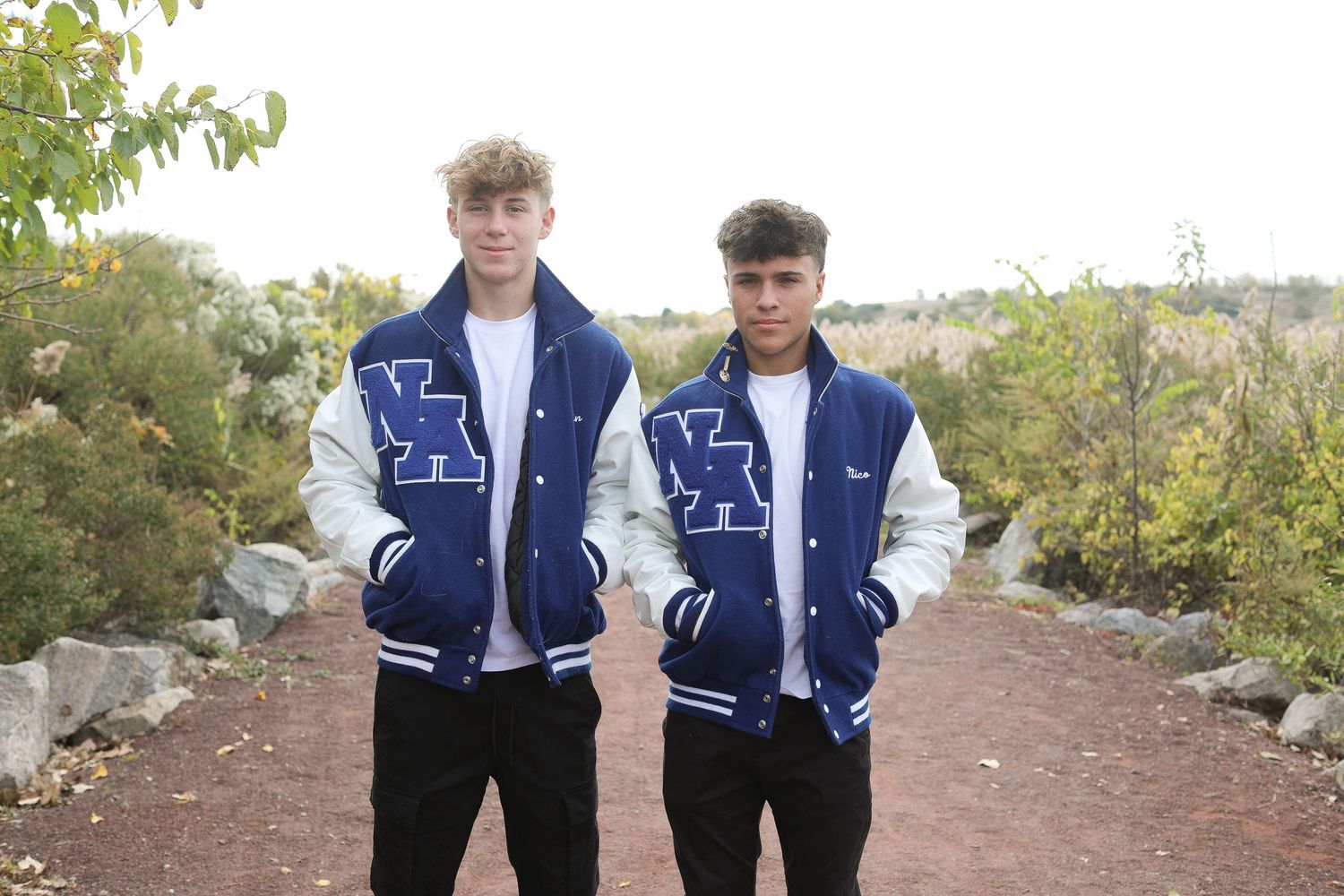Students wearing matching navy varsity jackets stand together on a pathway surrounded by greenery.