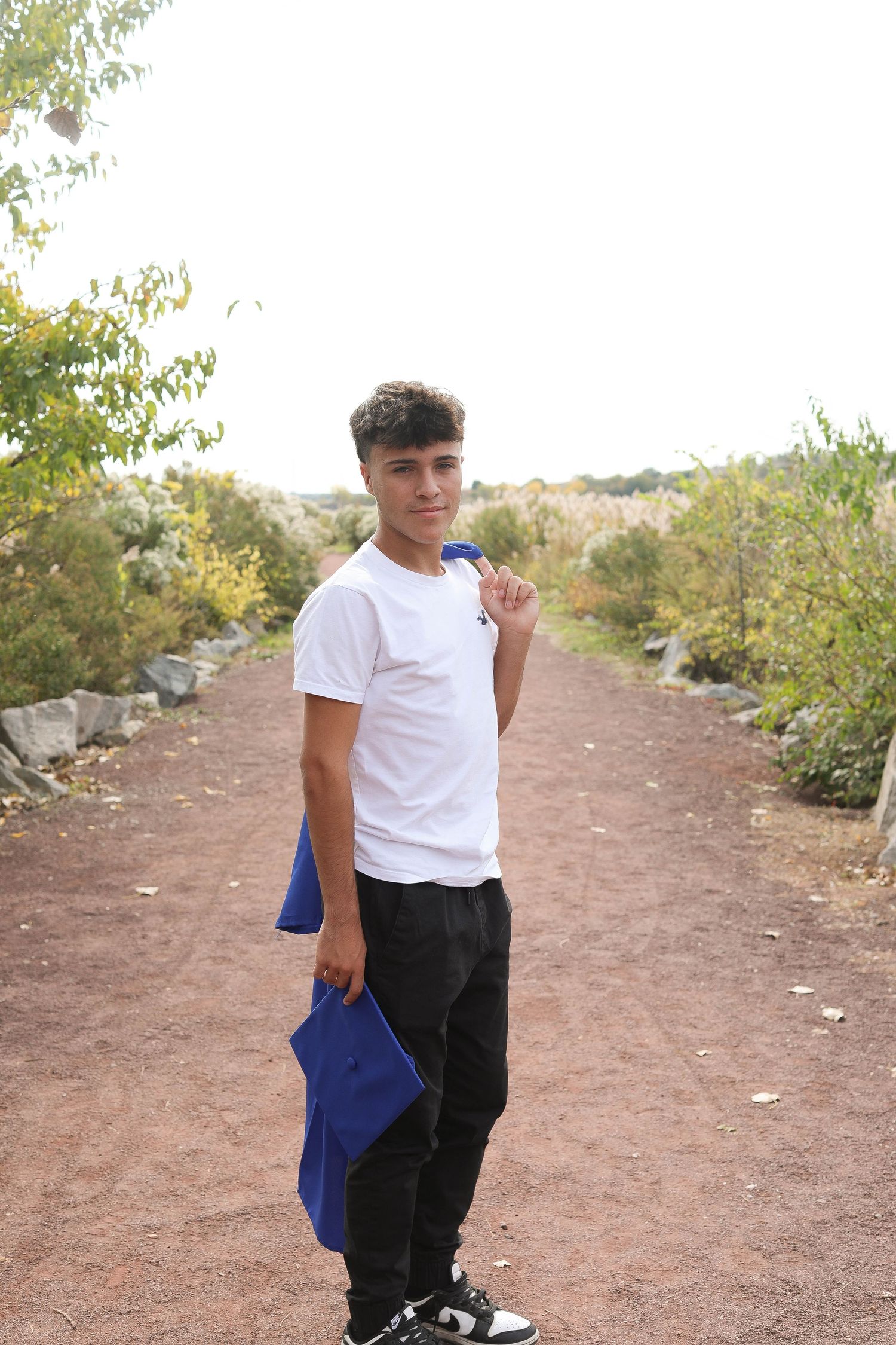 Person in casual attire holds blue graduation attire while standing on a nature trail.