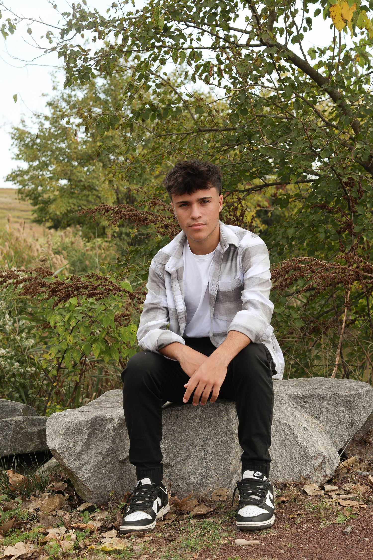 A casual outdoor portrait showing someone seated on a rock wearing a gray jacket, white t-shirt and black pants.