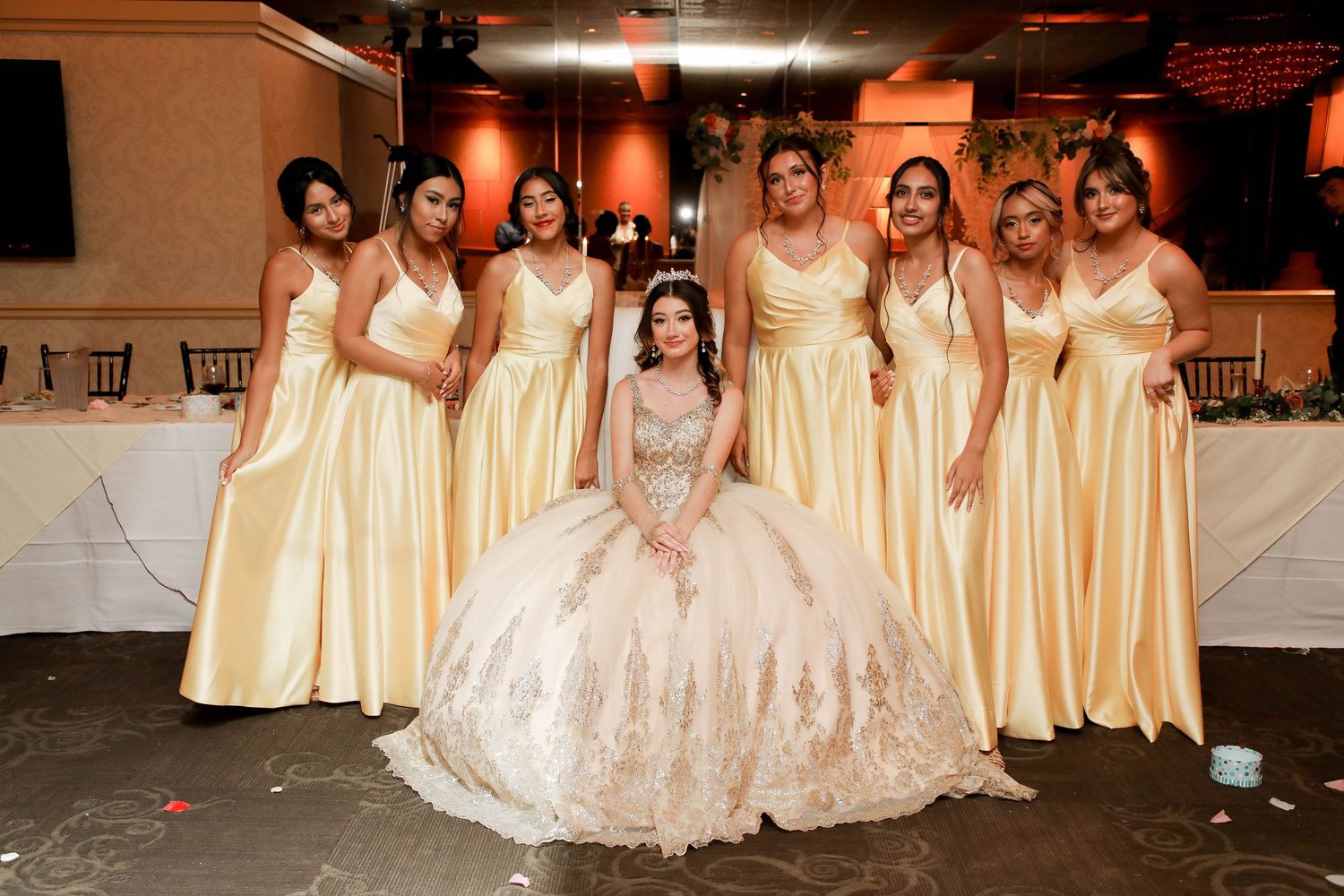 A bride in a sparkly ballgown sits surrounded by bridesmaids wearing matching yellow dresses at an indoor reception venue.