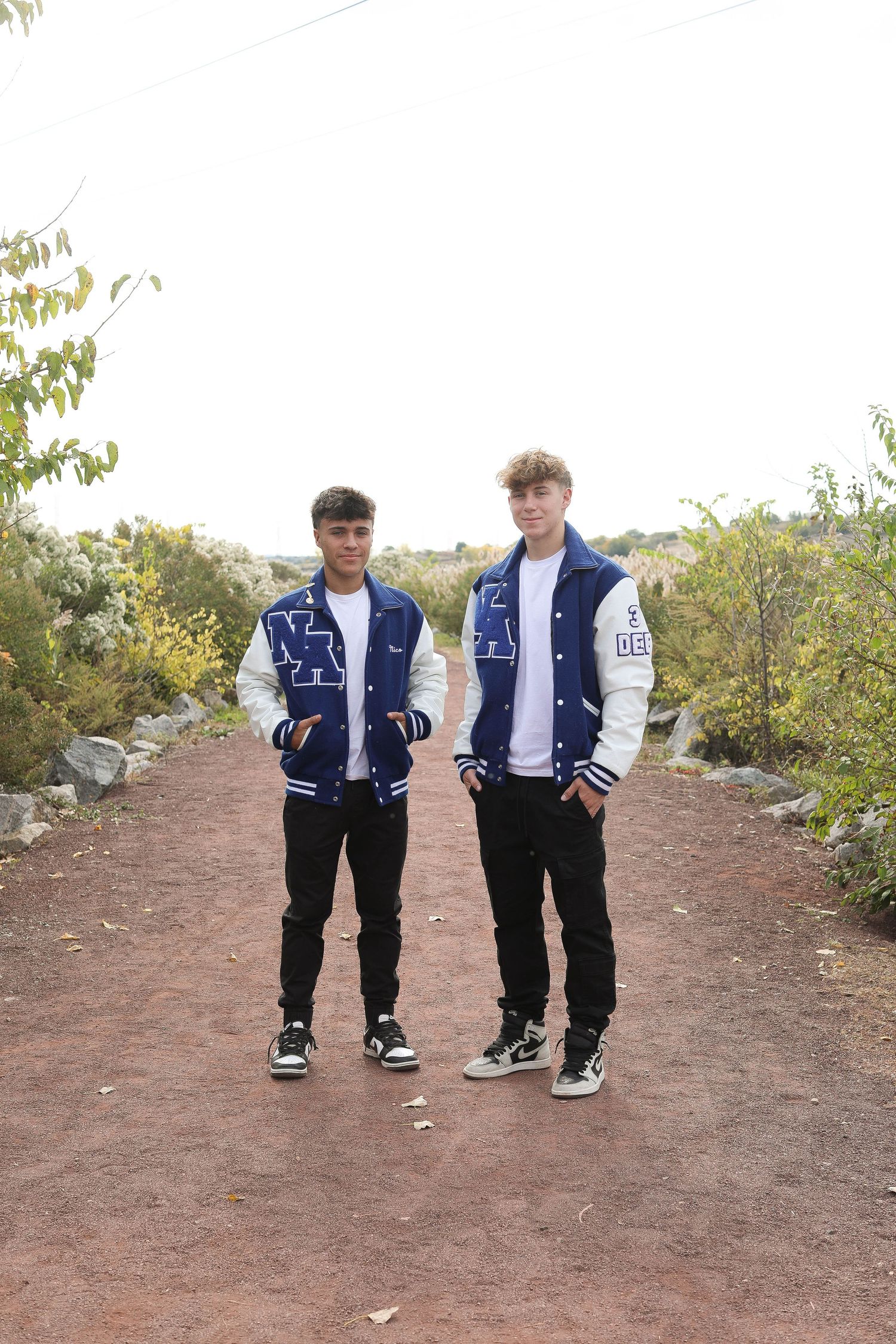 Two people wearing matching varsity jackets pose together on a dirt path.