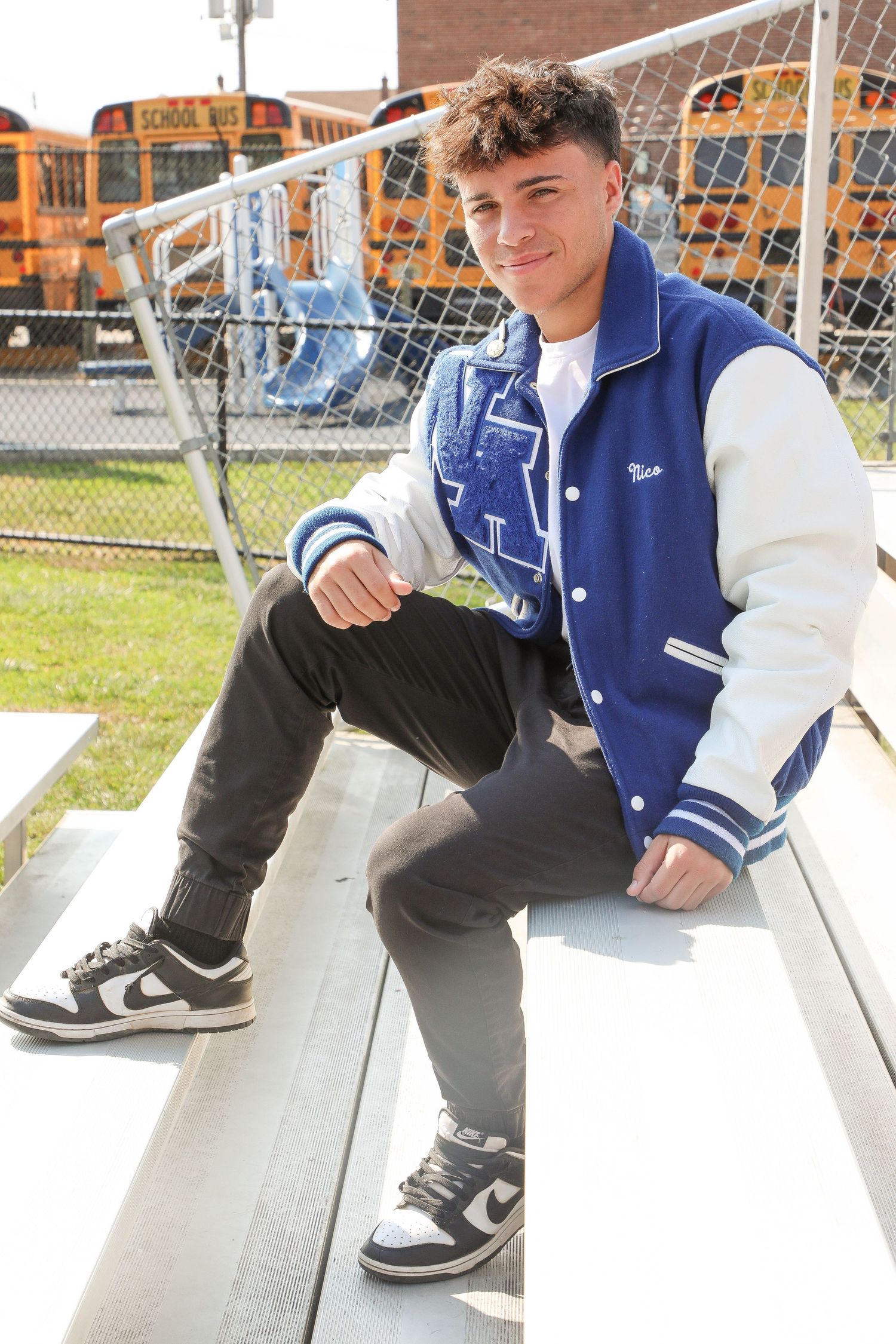 Athletic setting photo on bleachers wearing blue varsity jacket and sneakers.
