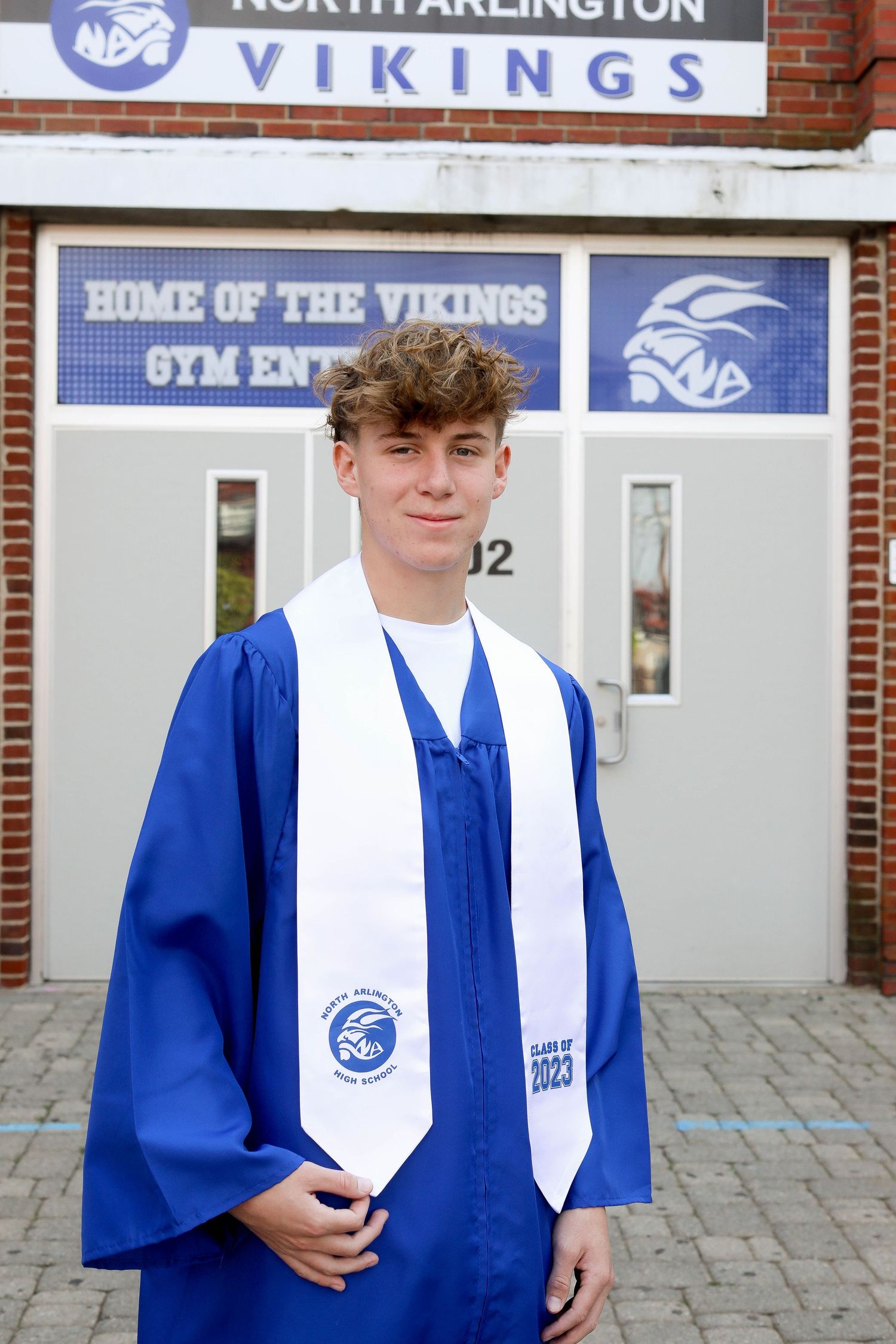 Graduate in blue cap and gown with white stole poses in front of Vikings gym entrance.