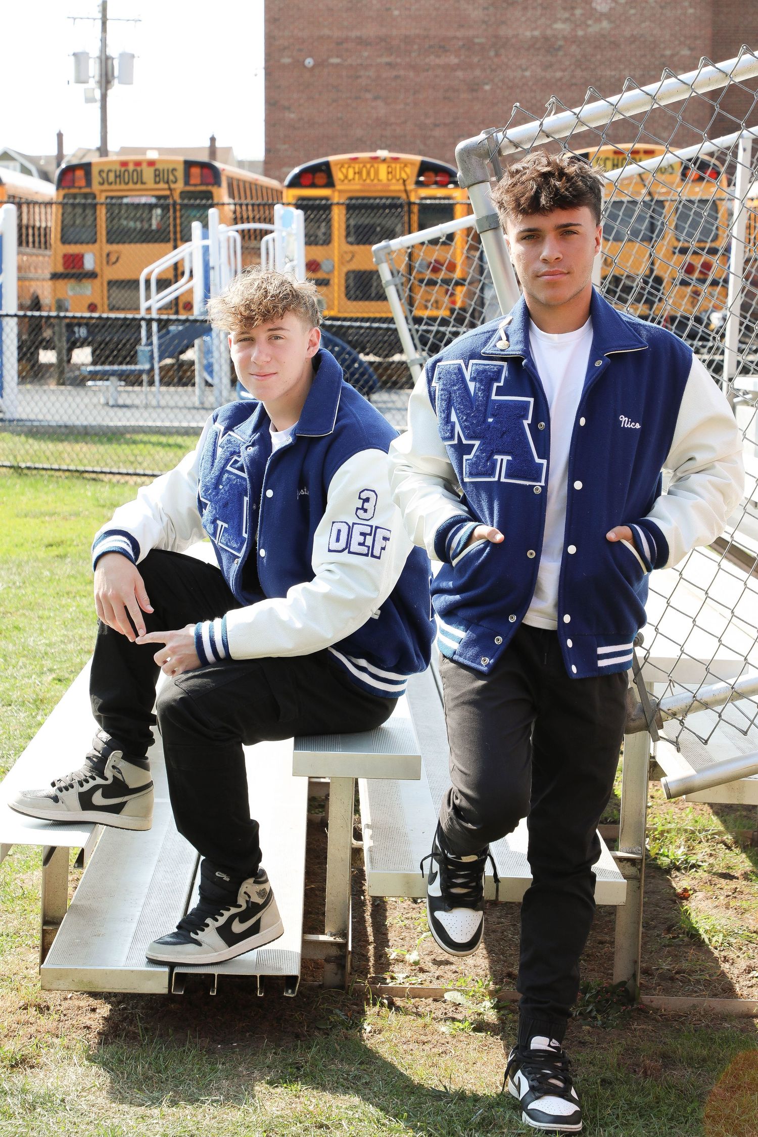 Two students wearing blue and white varsity jackets pose on bleachers with school buses in background.