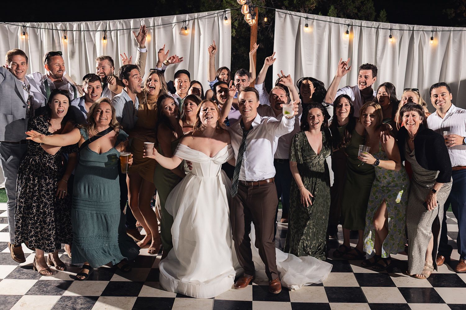 A festive wedding party celebrates on a black and white checkered dance floor under string lights at an indoor reception.