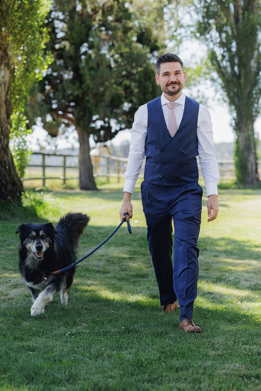 A person in a navy vest and dress pants walks a fluffy black and white dog on a leash in a grassy field.